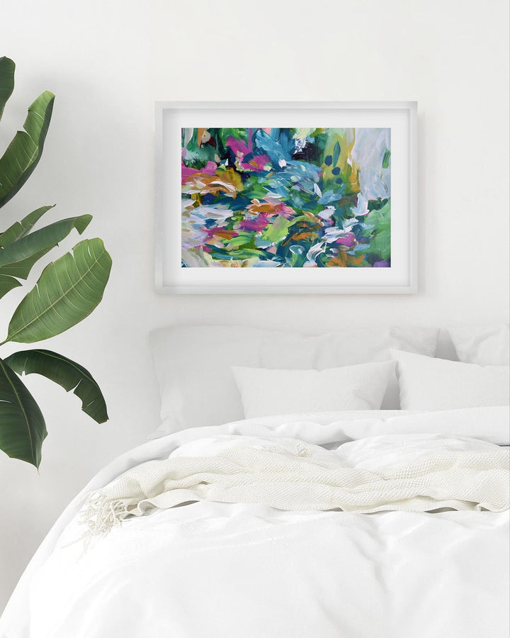 Framed abstract painting on a wall above a bed with a plant to the left.