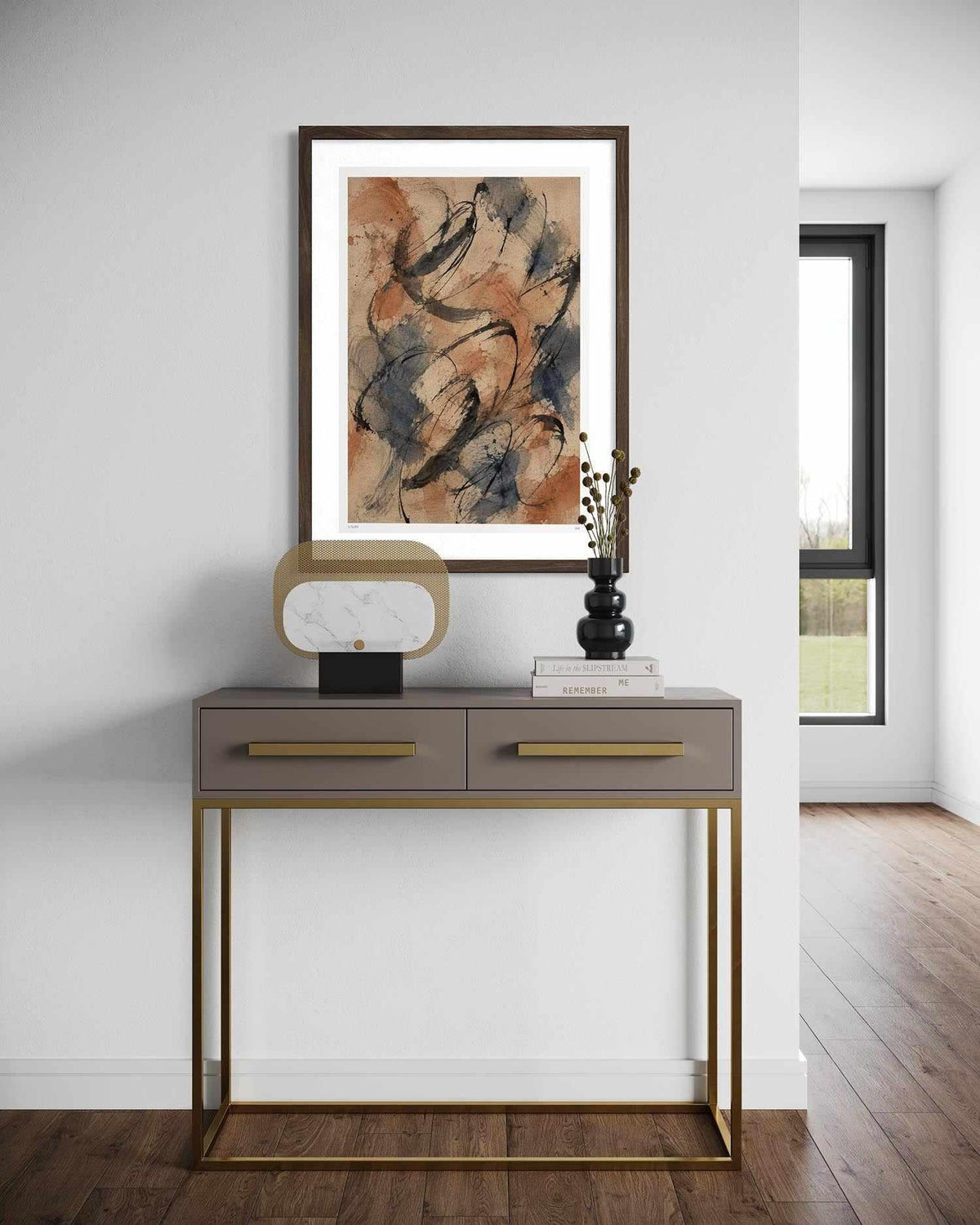 A framed abstract art print with warm earthy tones hangs on a white wall above a modern gray console table with gold-toned legs and handles. The room has wood flooring and a window in the background.