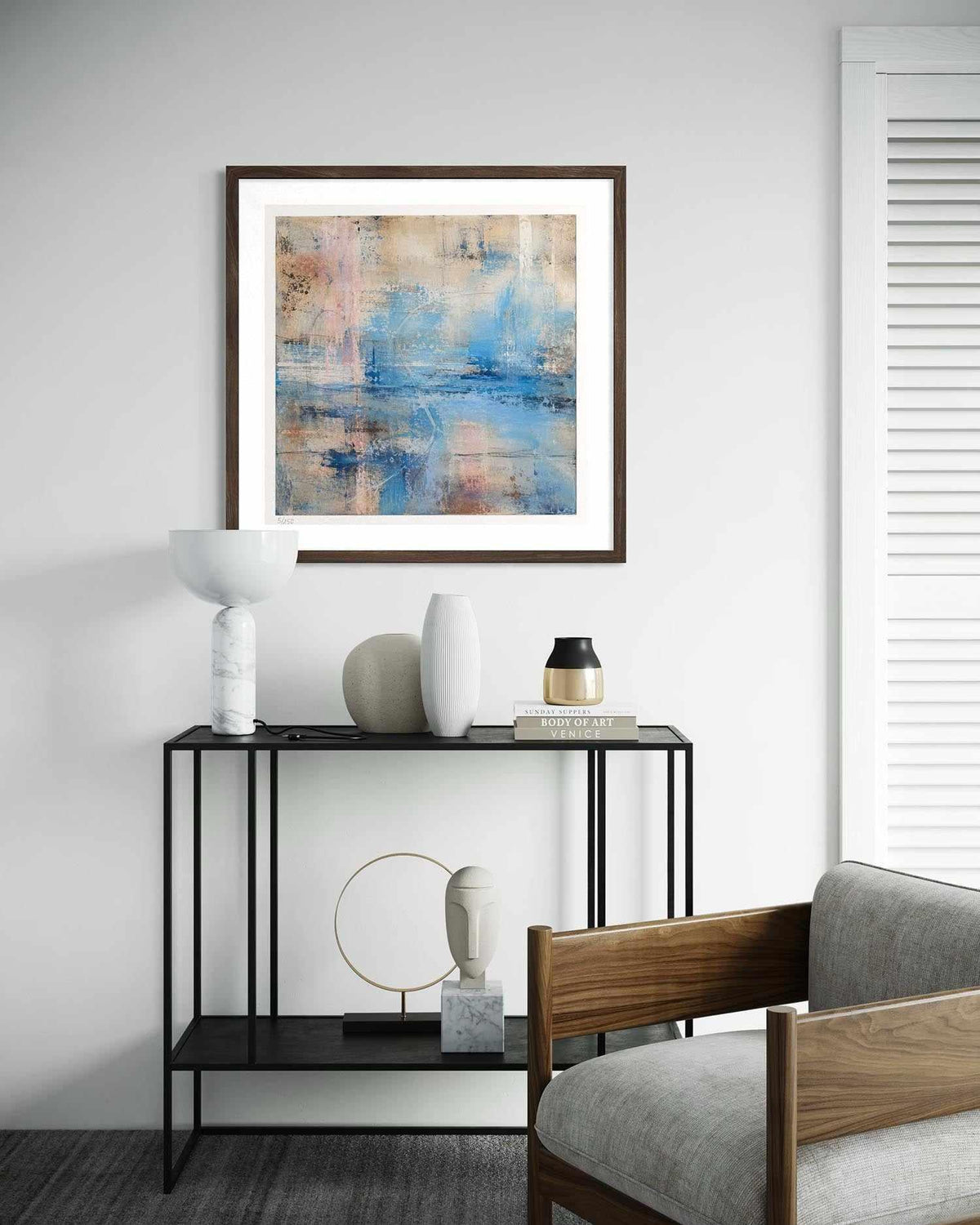 A framed abstract art print with blue, beige, and white tones hangs on a light gray wall above a black console table styled with decorative vases and a chair is partially visible in the foreground.