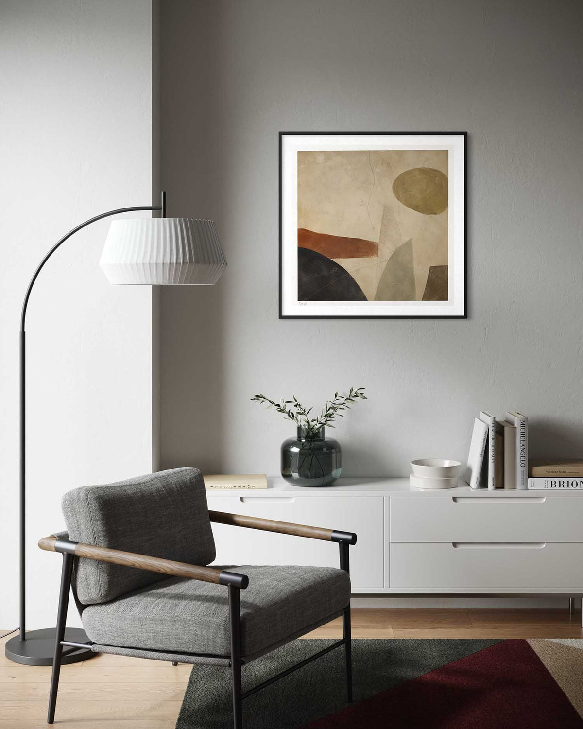 A framed abstract art print with beige, brown, and black geometric shapes hangs on a gray wall in a modern living room setting above a white sideboard, next to a gray armchair and a black floor lamp.