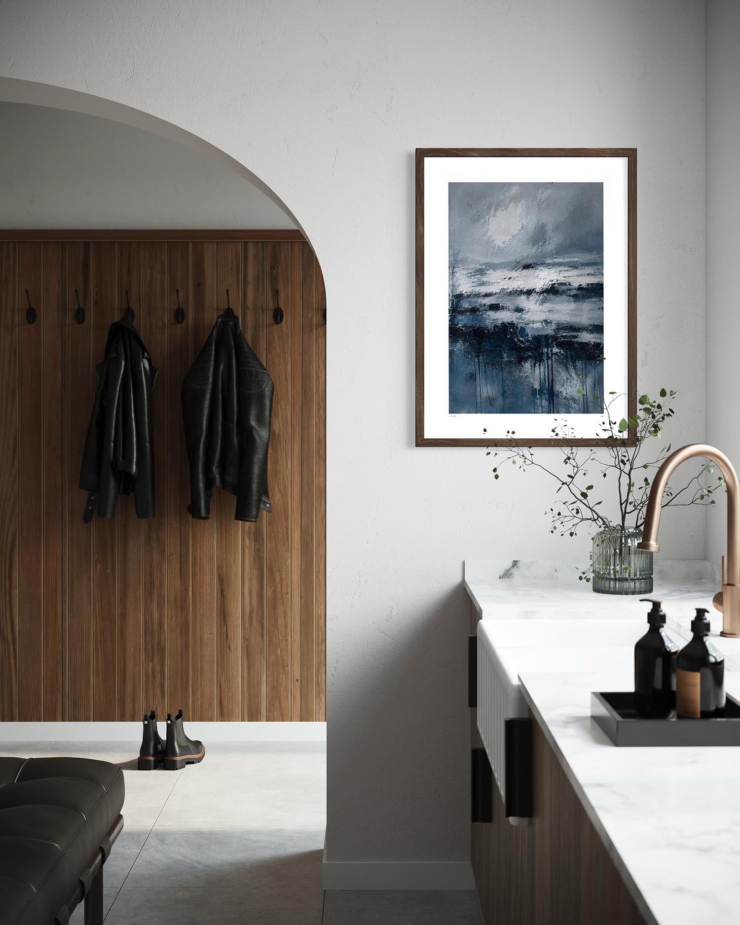 A framed square art print featuring a moody blue and gray seascape hangs on a white wall in a modern kitchen entryway with a wood-paneled door, hanging jackets, and a marble countertop.