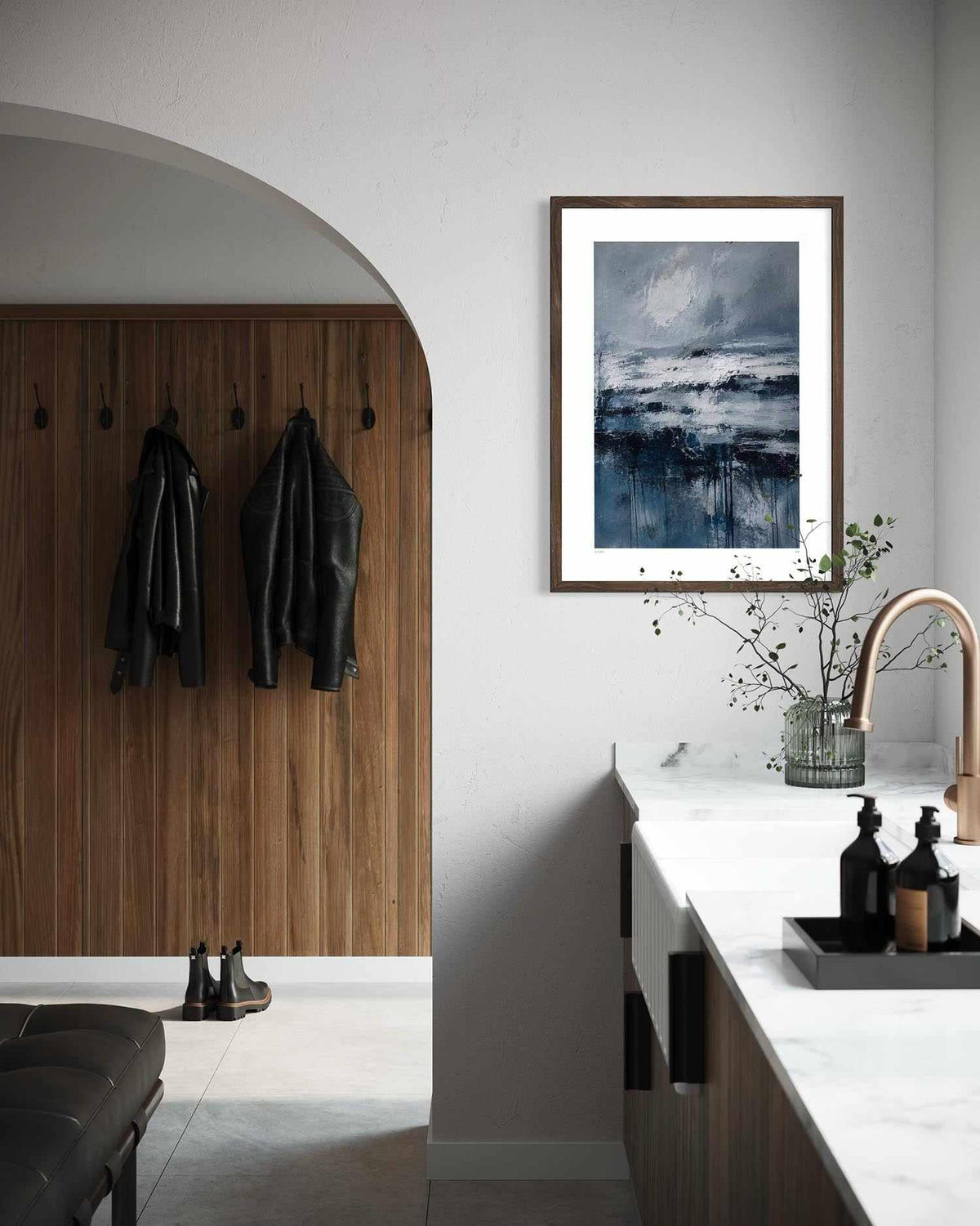 A framed square art print featuring a moody blue and gray seascape hangs on a white wall in a modern kitchen entryway with a wood-paneled door, hanging jackets, and a marble countertop.