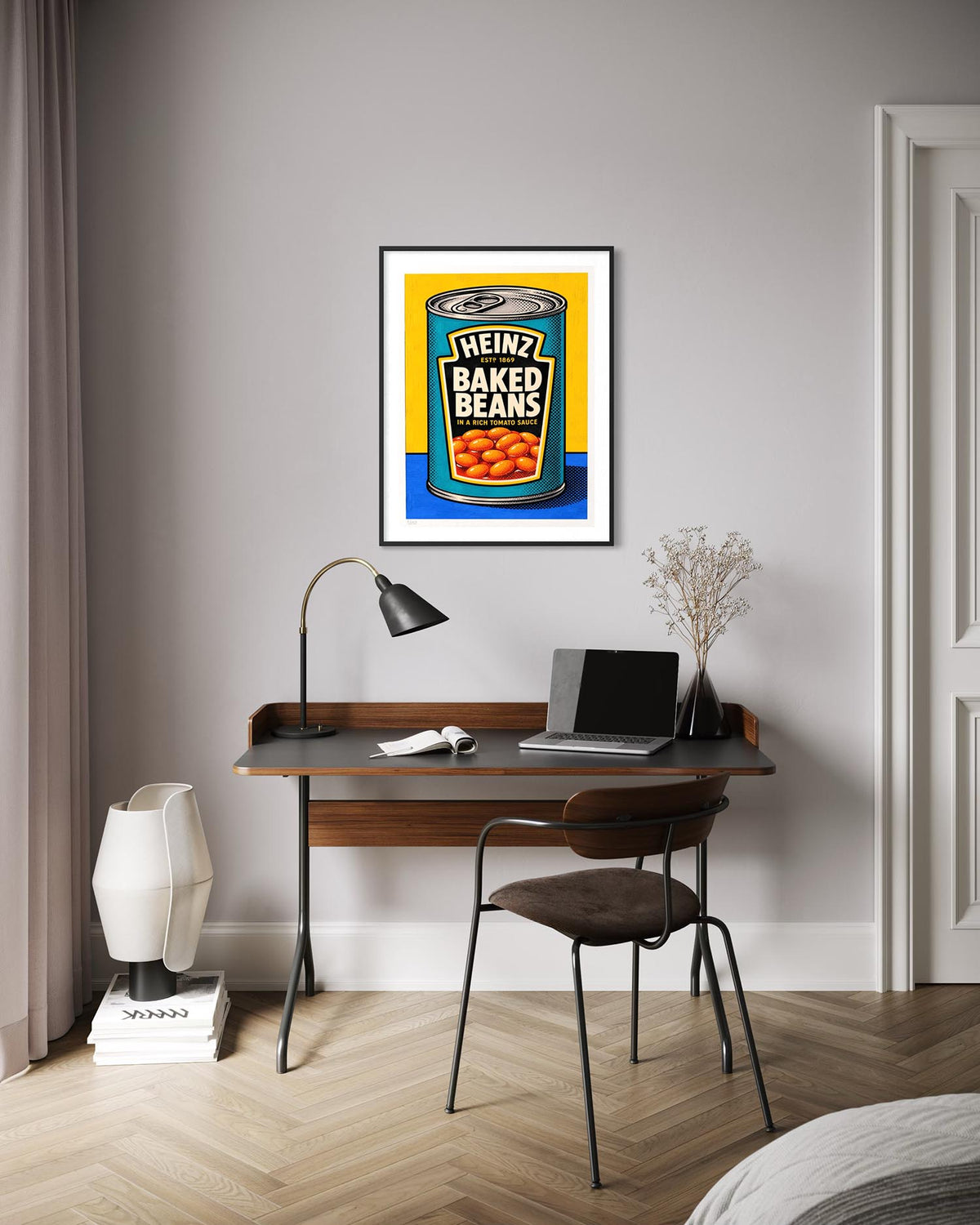 A framed pop art print of a Heinz Baked Beans can hangs on a light gray wall above a wooden desk with a laptop, lamp, and chair in a modern room setting.