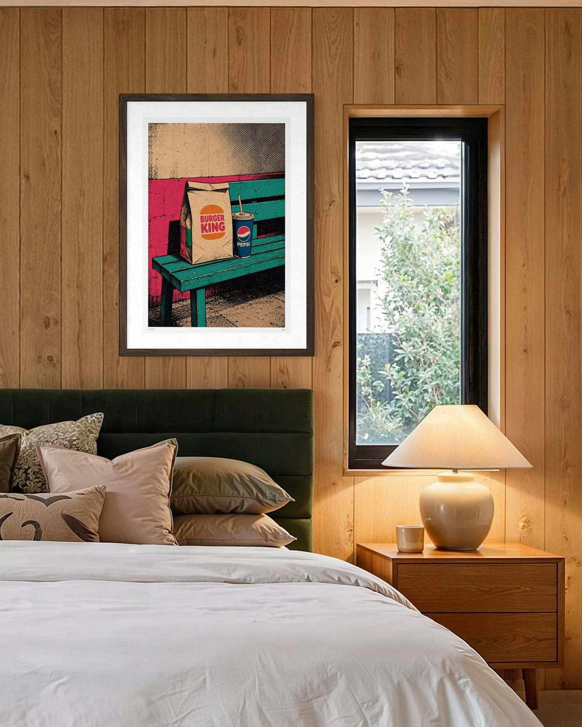 A framed art print featuring a colorful scene of a green bench with a Burger King bag and a Pepsi cup, displayed on a wood-paneled wall above a bed in a bedroom setting.