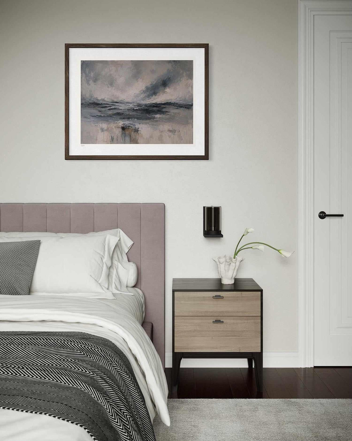 A framed abstract landscape art print with a dark wood frame and white mat, hanging on a light gray wall above a bed in a modern bedroom setting.
