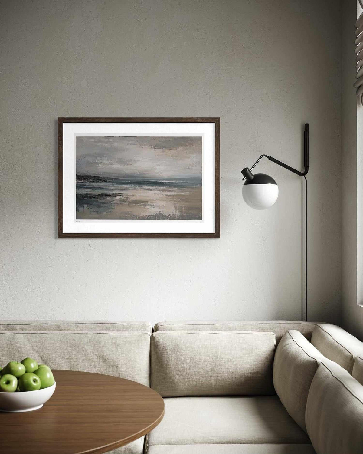 A framed landscape art print with a dark brown frame and white mat, hanging on a textured gray wall above a beige sofa in a modern living room setting.
