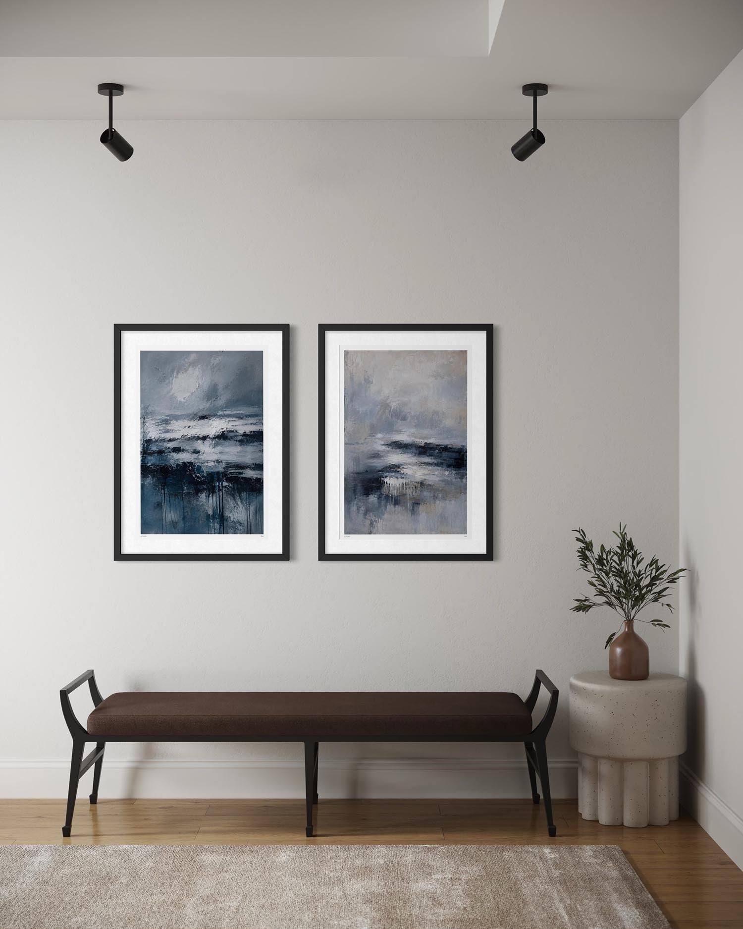 Two framed abstract landscape art prints with black frames and white mats hang on a light gray wall above a brown upholstered bench. A small side table with a plant sits to the right, and a rug covers the wooden floor.