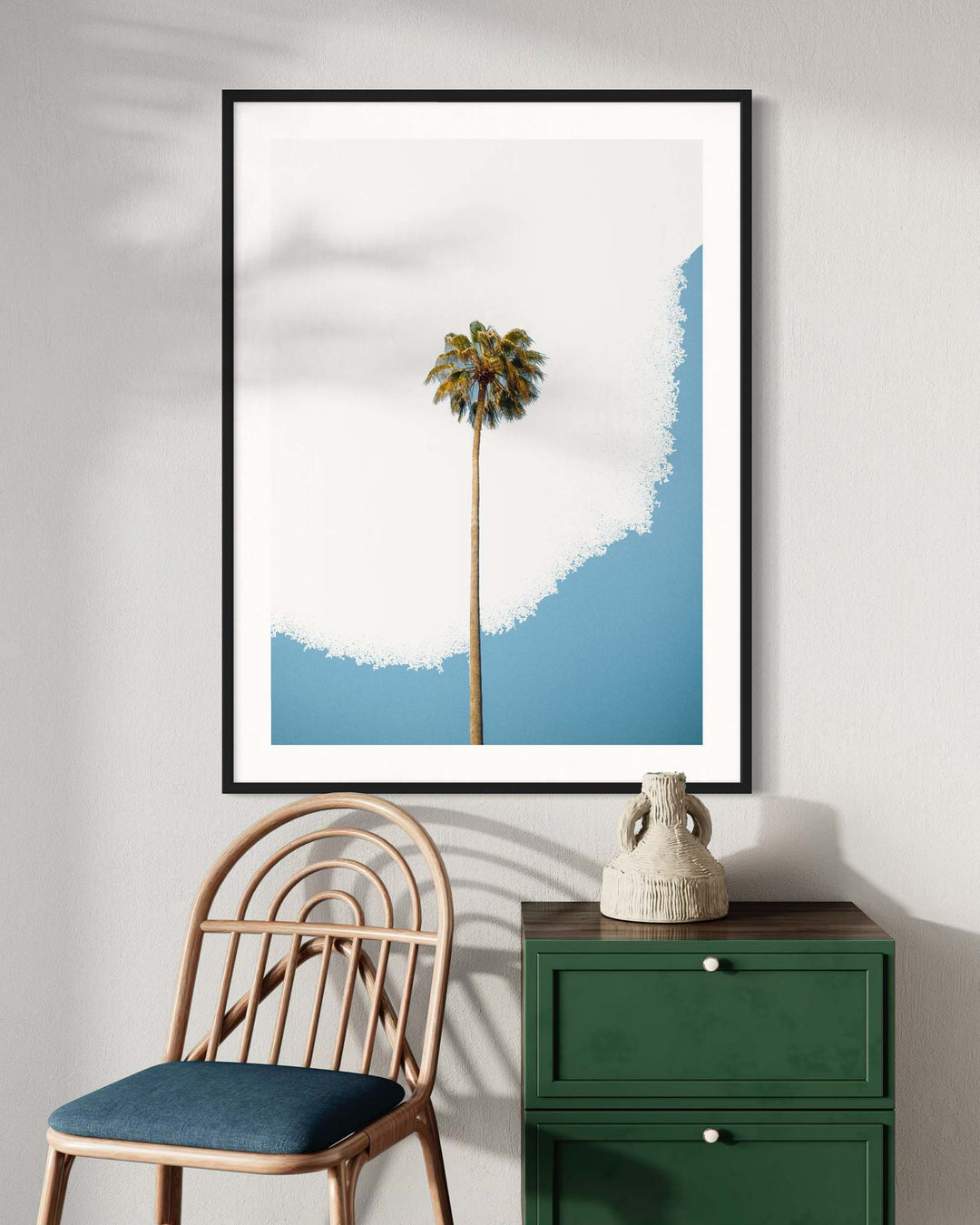 Framed artwork of a palm tree with a blue and white gradient on a wall above a chair and green cabinet.