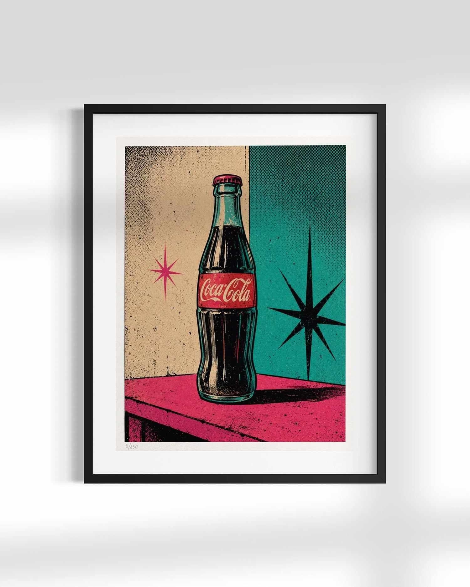 A framed retro-style Coca-Cola art print featuring a glass bottle on a pink table against a split beige and teal background with starburst accents, shown hanging on a light gray wall.