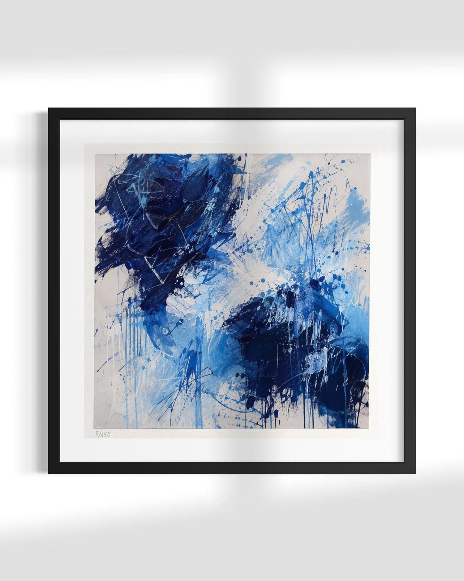 Framed abstract wall art featuring expressive blue and navy brushstrokes and splatters on a white and light gray background, presented in a black frame with a white mat against a light gray wall.