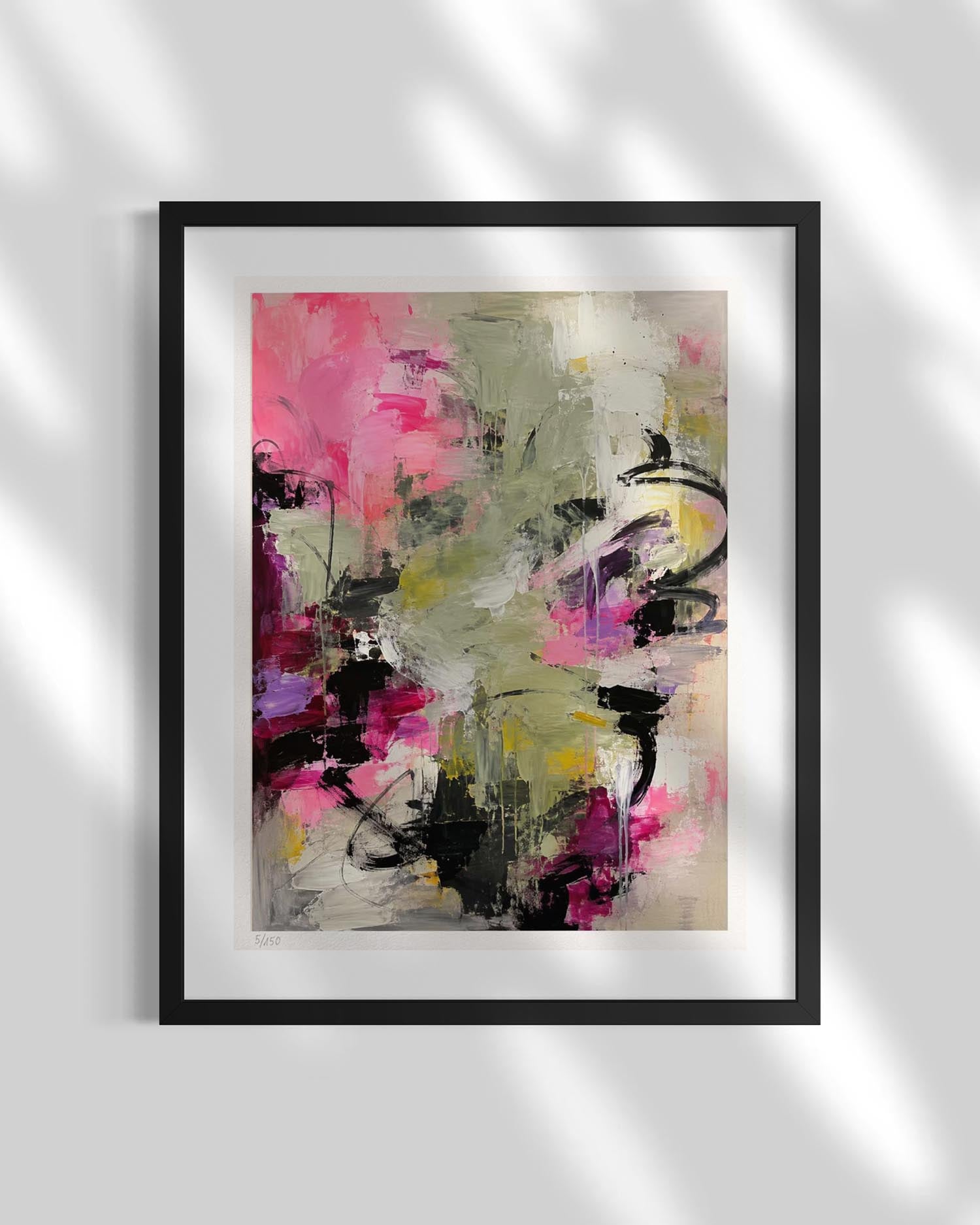 A framed abstract art print featuring expressive brushstrokes in pink, olive green, black, purple, and yellow on a white mat, set in a black frame against a light gray wall with soft leaf shadows.