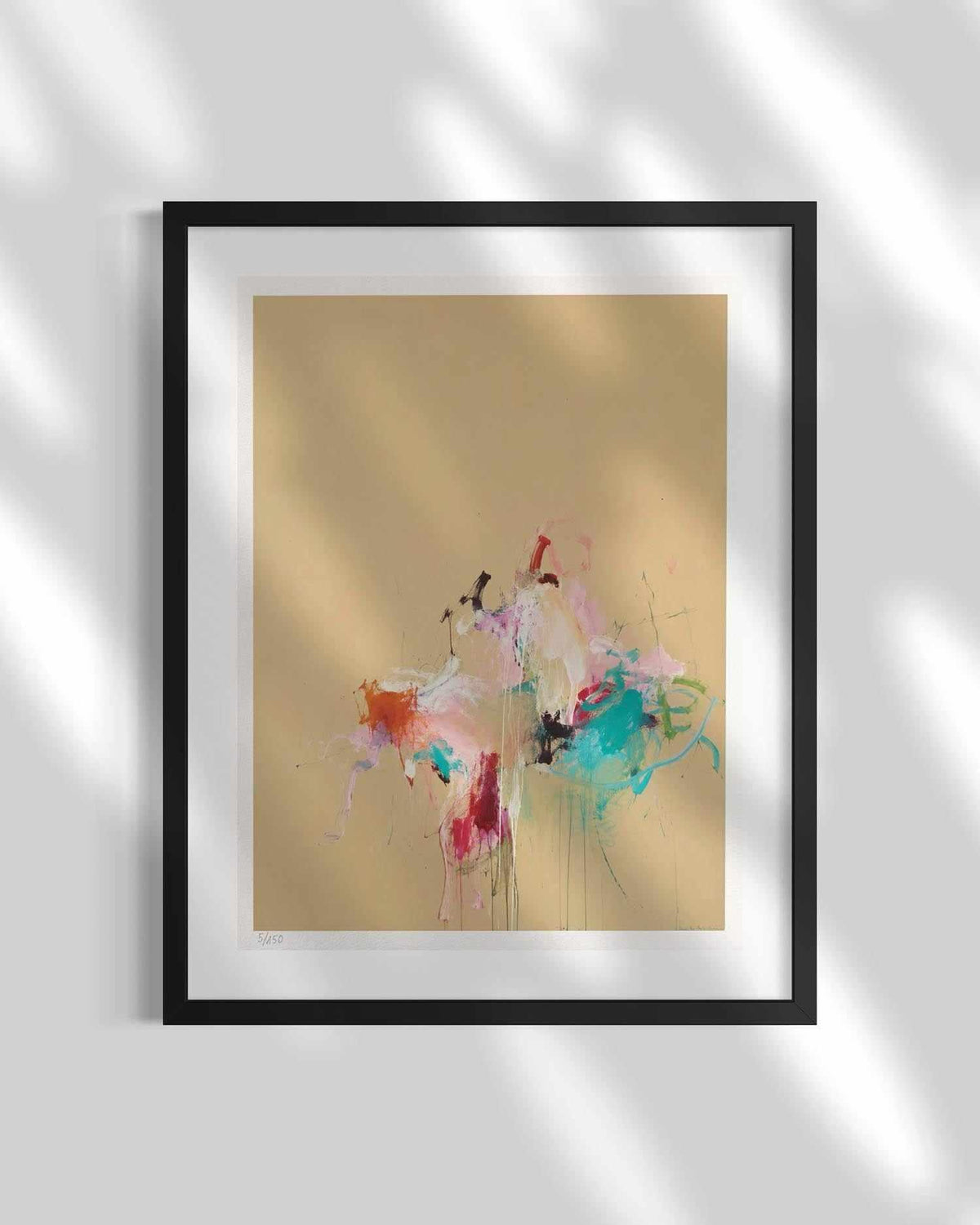 Framed abstract wall art hanging on a light gray wall, with soft leaf shadows cast across the print. The artwork features a warm beige background with colorful paint splashes and drips in pink, white, blue, red, and orange, set within a black frame and white mat.