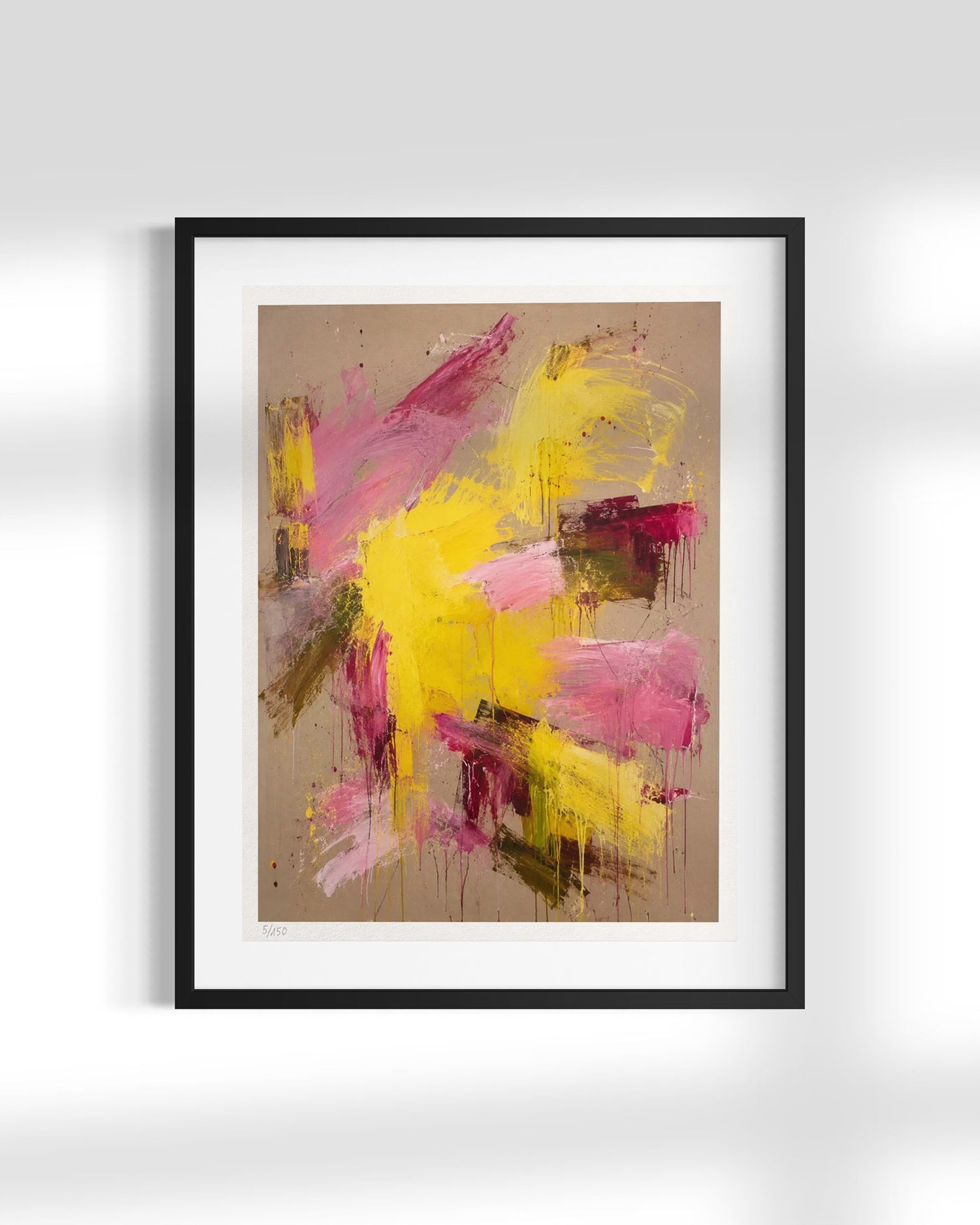 A framed abstract art print featuring expressive brushstrokes in yellow, pink, and magenta on a warm beige background, enclosed in a black frame with a white mat, shown against a light gray wall.