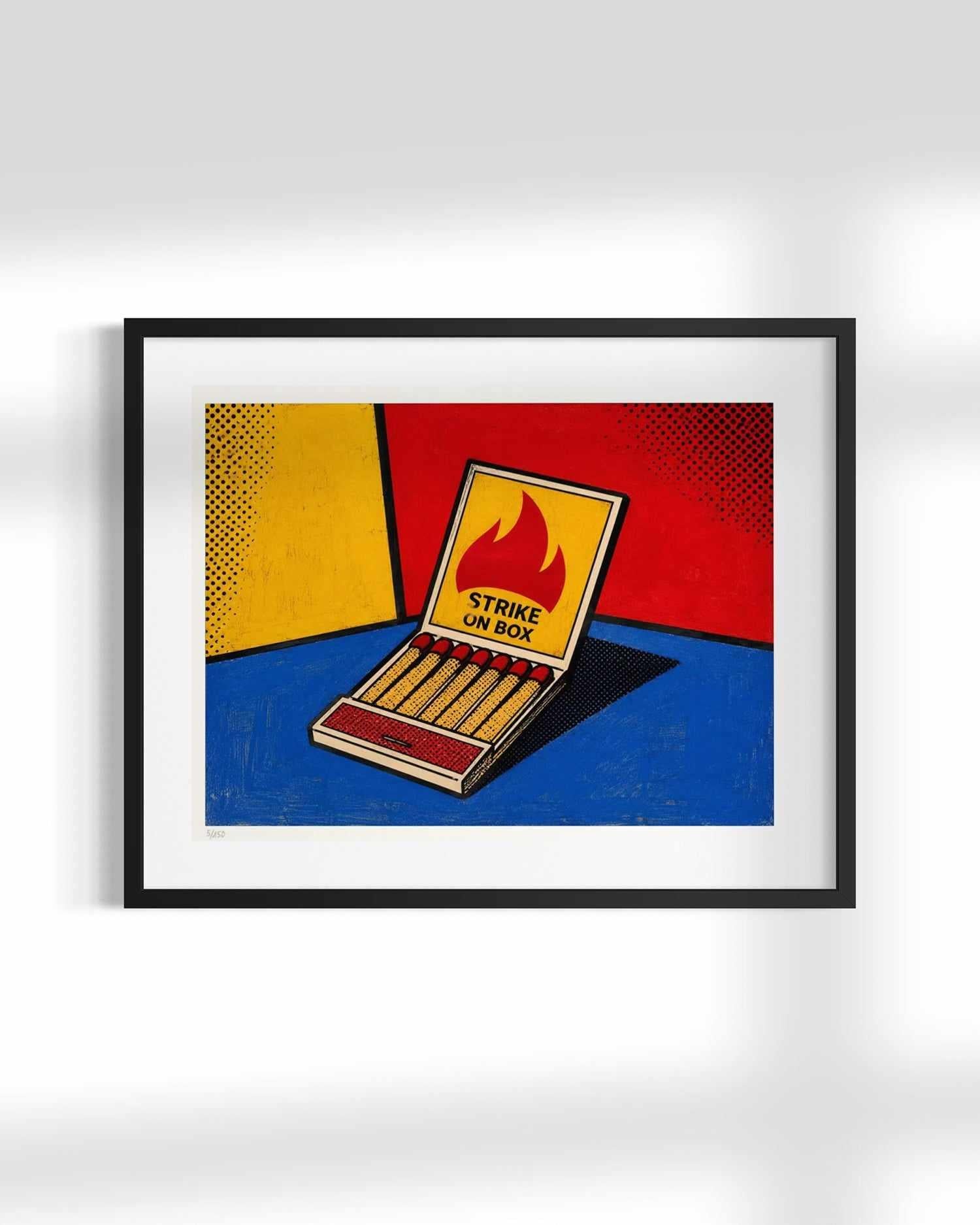 Framed pop art print featuring a matchbox with the words 'STRIKE UN BOX' and a flame graphic, set against a bold red, yellow, and blue background, displayed on a light gray wall.