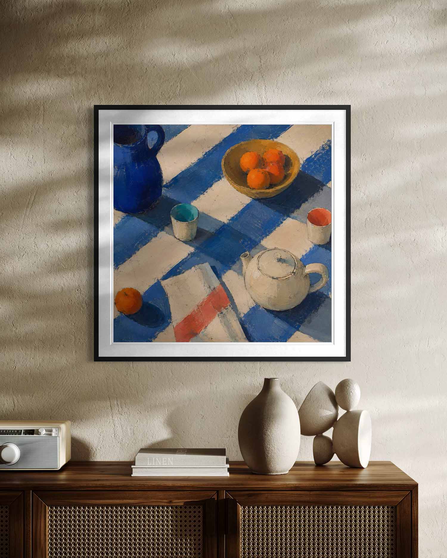 A framed still life painting hanging on a textured beige wall above a wooden cabinet. The artwork depicts a blue pitcher, a bowl of oranges, a teapot, and small cups on a blue-and-white striped cloth.