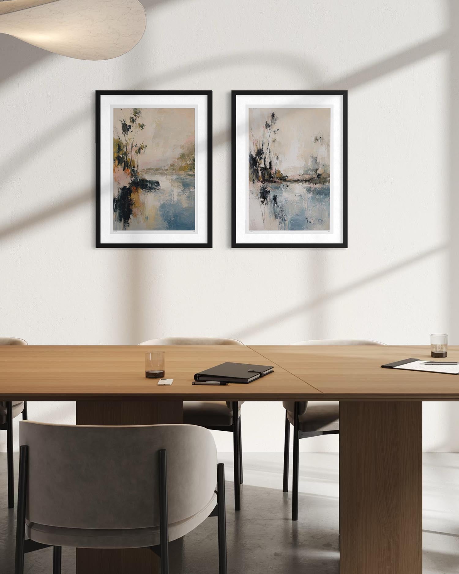 Two framed abstract landscape art prints with black frames and white mats, hanging side-by-side on a white wall above a wooden conference table and gray chairs.