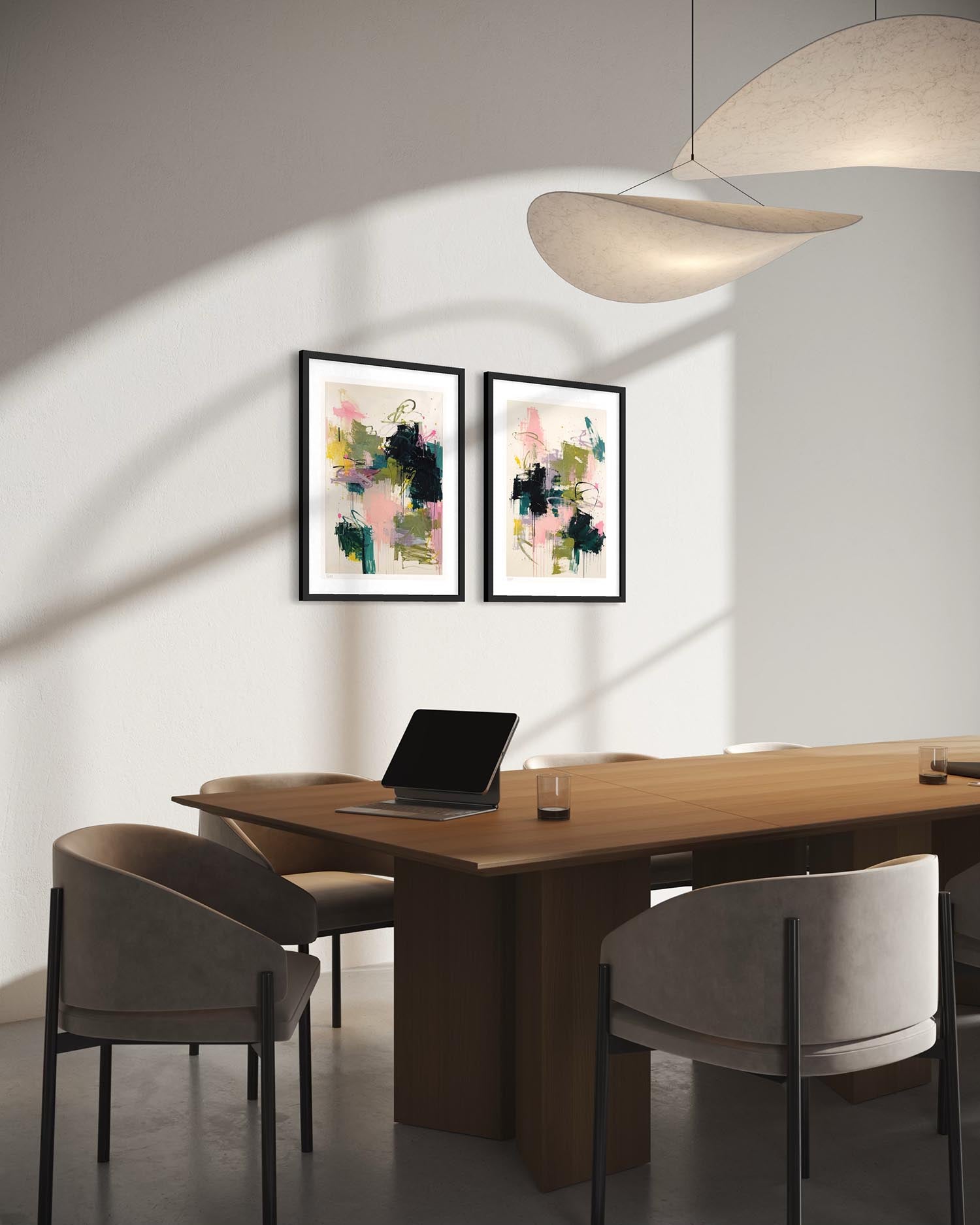 Two framed abstract art prints with black frames hang on a light gray wall above a wooden dining table with gray upholstered chairs in a modern, sunlit room.