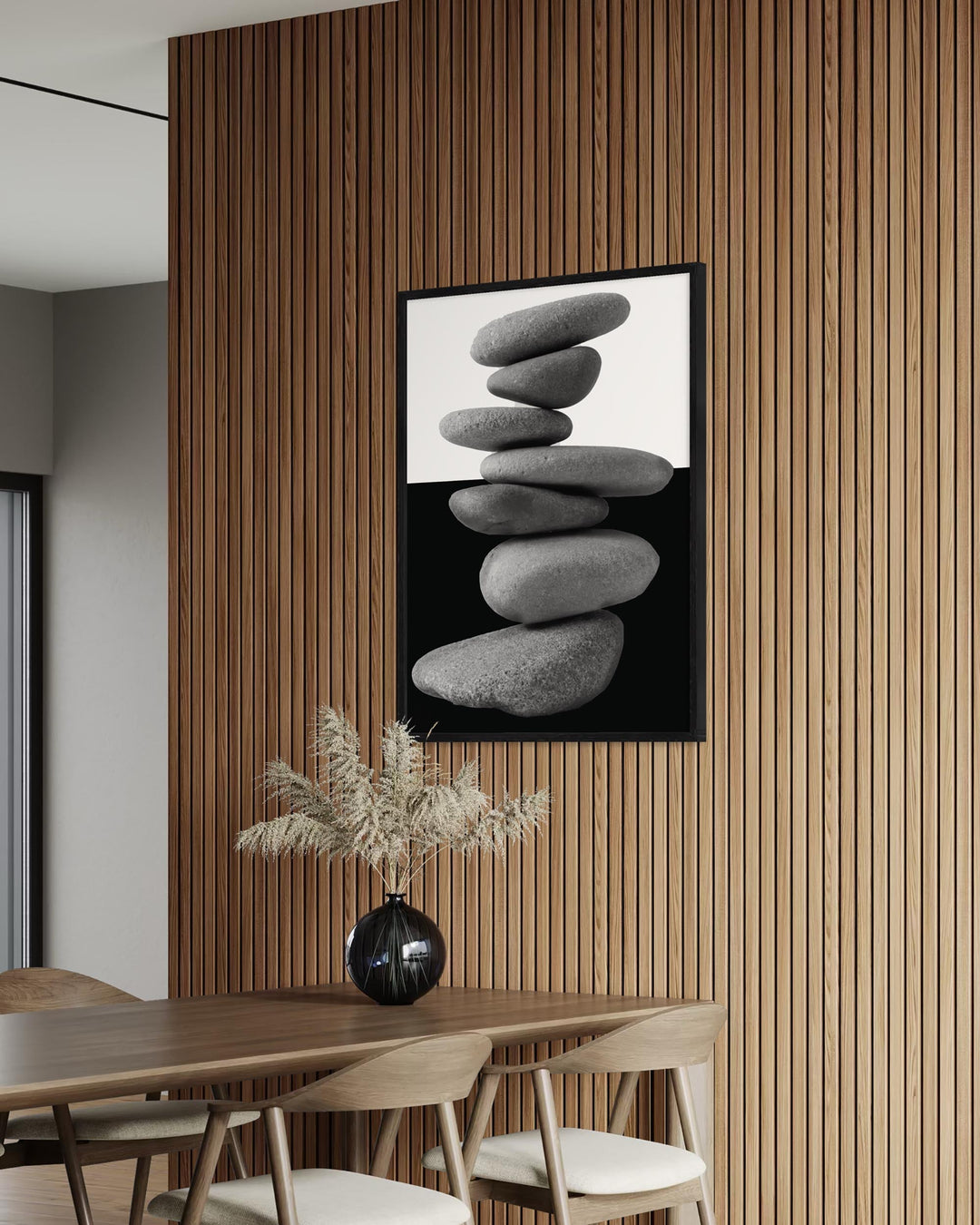 Dining room with a wooden wall, framed artwork of stacked stones, and a vase with dried plants on a table.