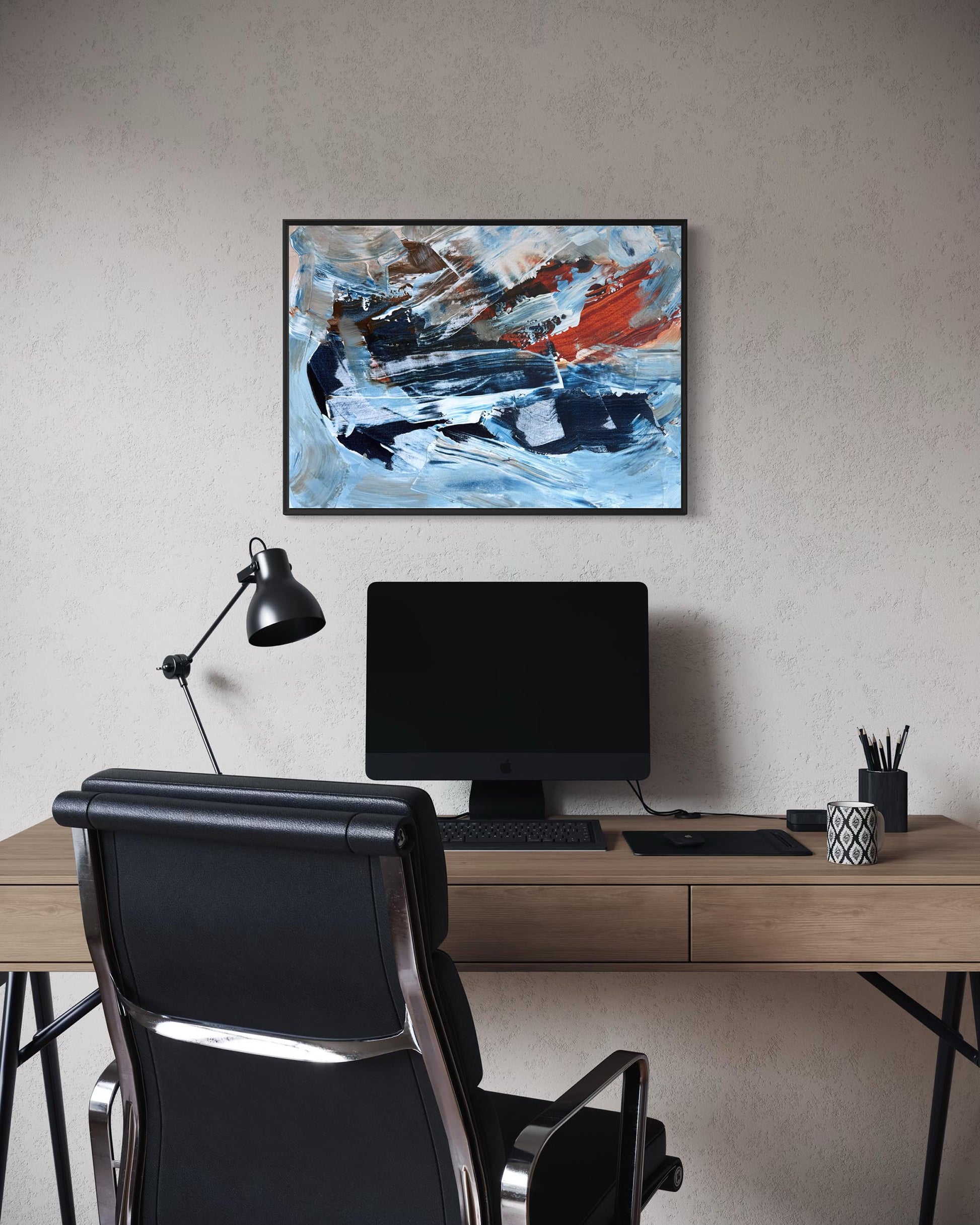 An abstract framed painting with blue, white, black, and red brushstrokes hangs on a textured beige wall above a modern desk setup with a computer monitor, black desk lamp, and black office chair.