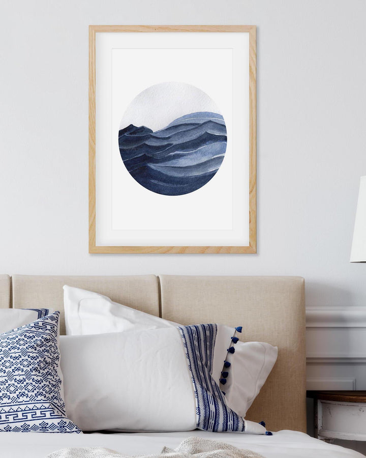 Framed abstract art of wavy blue design on a white wall above a bed with decorative pillows.