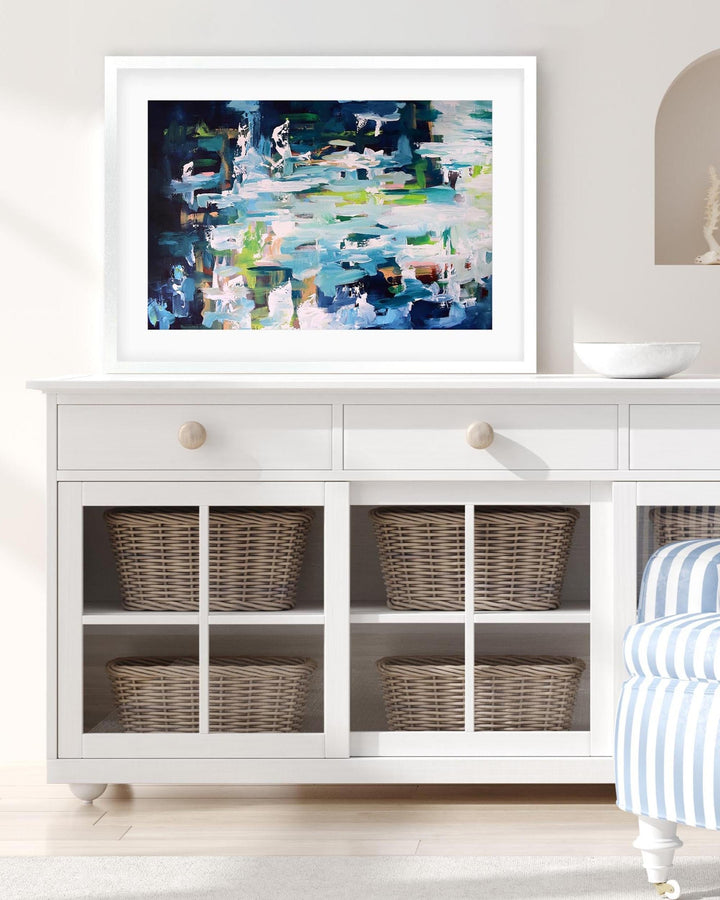 Framed abstract painting on a white wall above a white cabinet with wicker baskets.