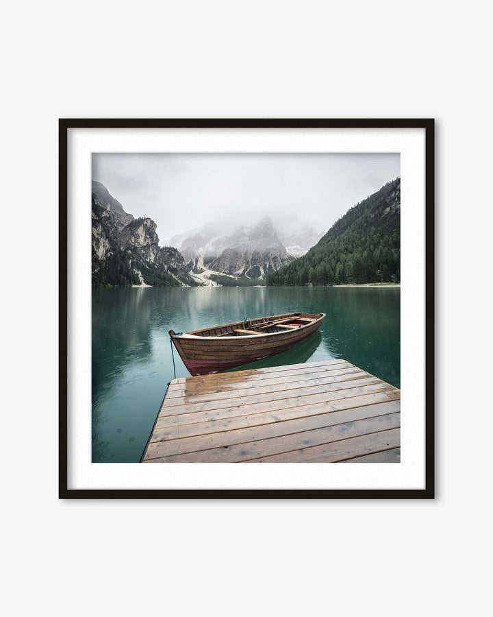 Framed photograph of a wooden boat on a dock with mountains and a lake in the background.