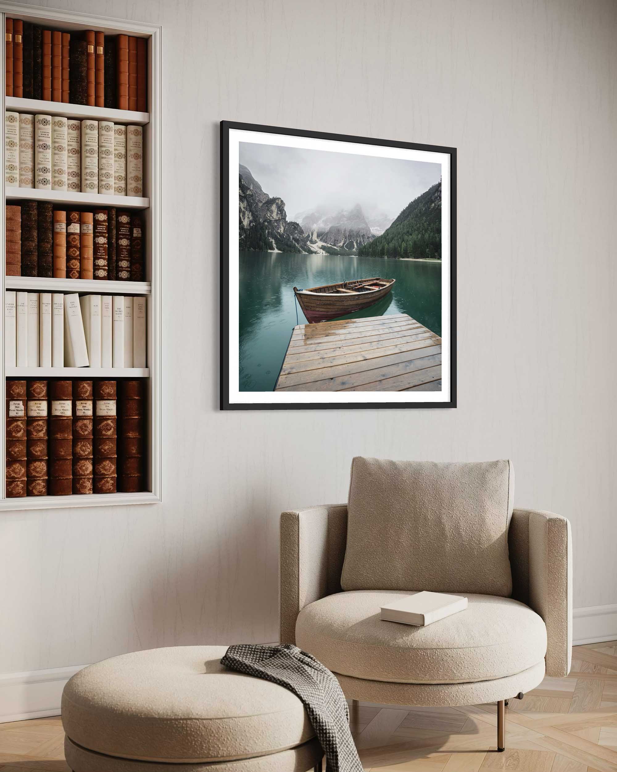 Framed landscape print of a lake with a boat on a wooden dock, above a beige armchair with a book.