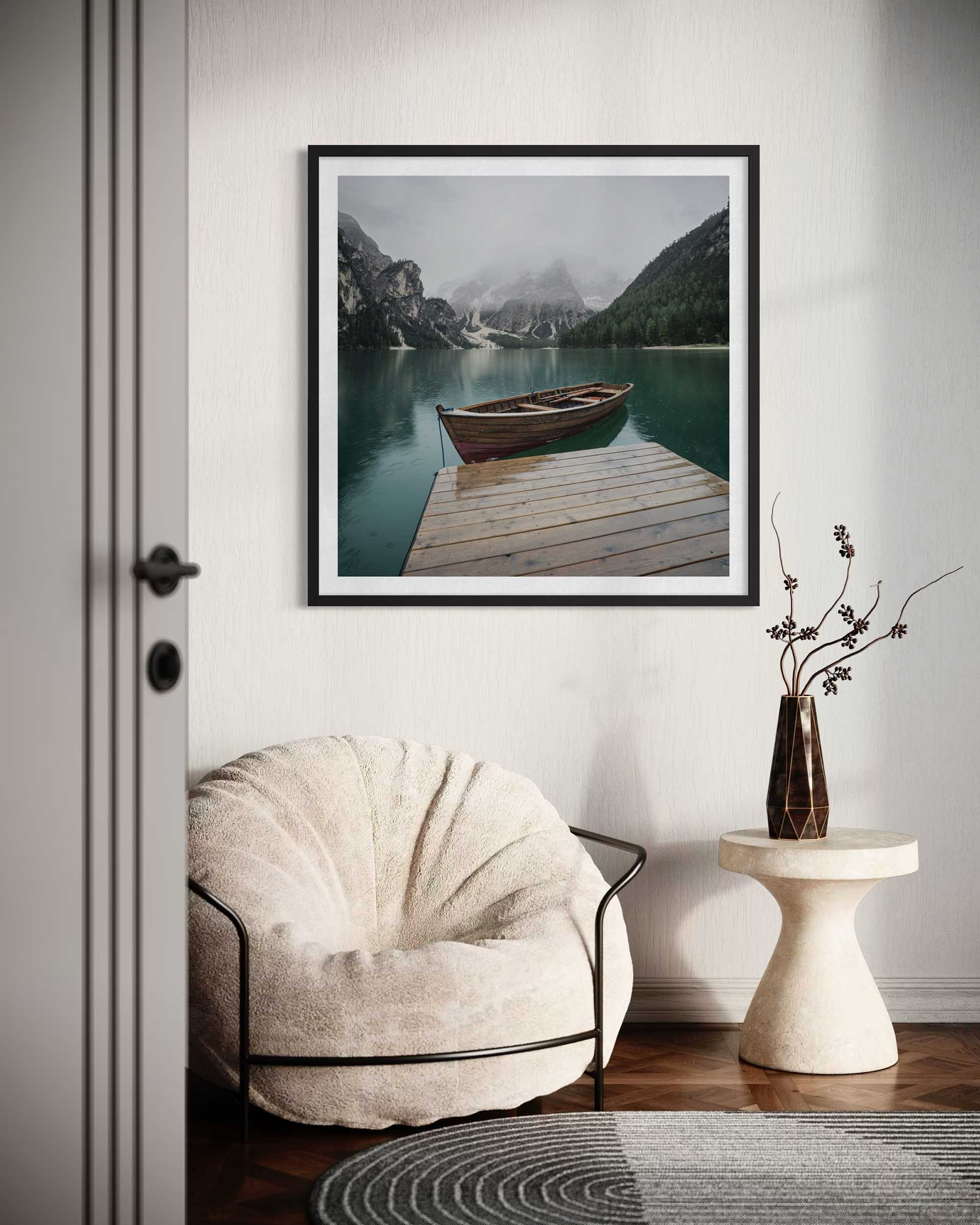 Framed landscape print of a lake with a boat on a wooden dock, above a beige armchair in a room.