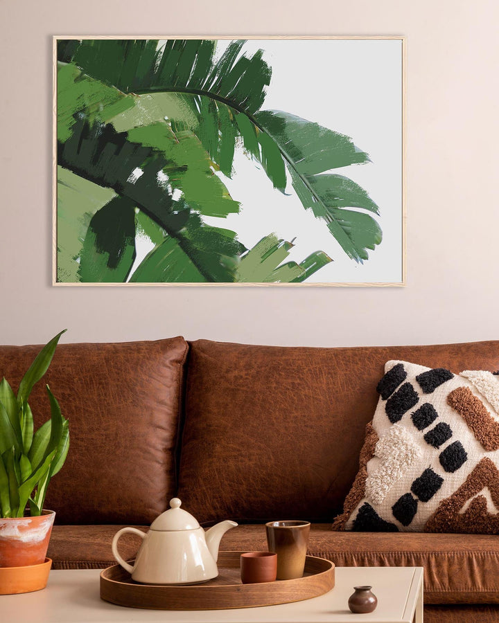 Framed artwork of green leaves above a brown sofa with decorative items on a table.