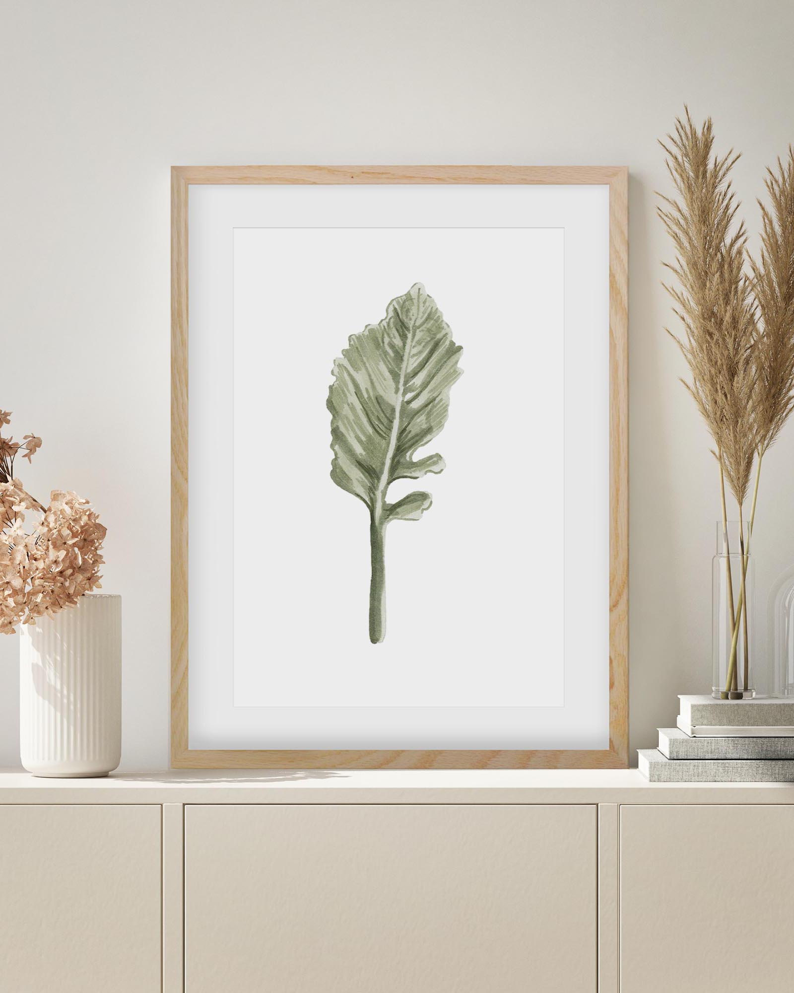 Framed botanical print of a green leaf on a white background, displayed on a shelf with decorative items.
