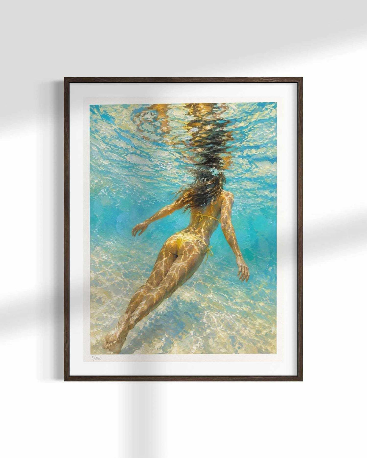 A framed art print featuring an underwater photograph of a woman swimming, displayed on a white wall with soft shadows.