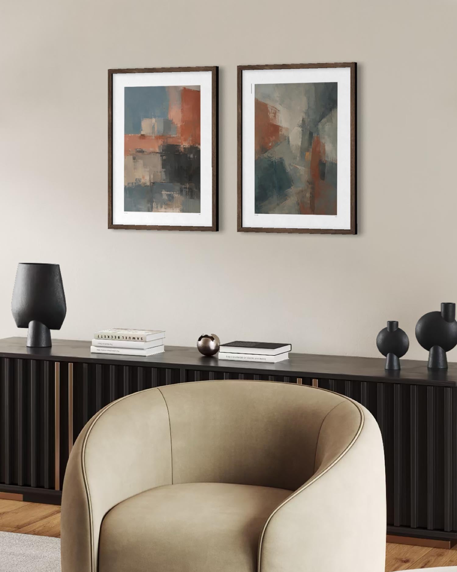 Two framed abstract wall art prints hang above a dark console table in a styled interior. The room includes a beige upholstered armchair in the foreground, black sculptural lamps, books, and a light wood floor.