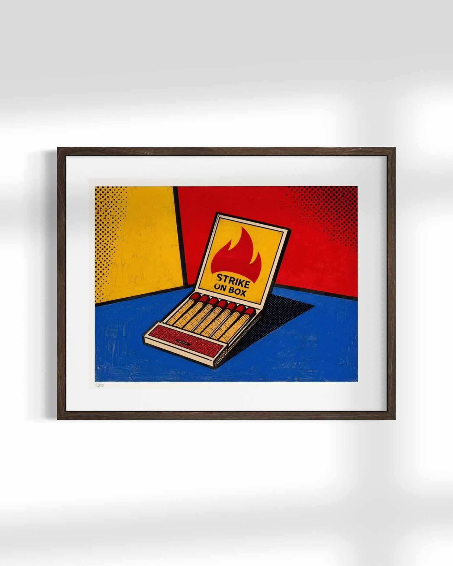 Framed pop art print of an open matchbox with a flame label reading 'STRIKE ON BOX', set against a bold yellow, red, and blue background, displayed on a light gray wall.