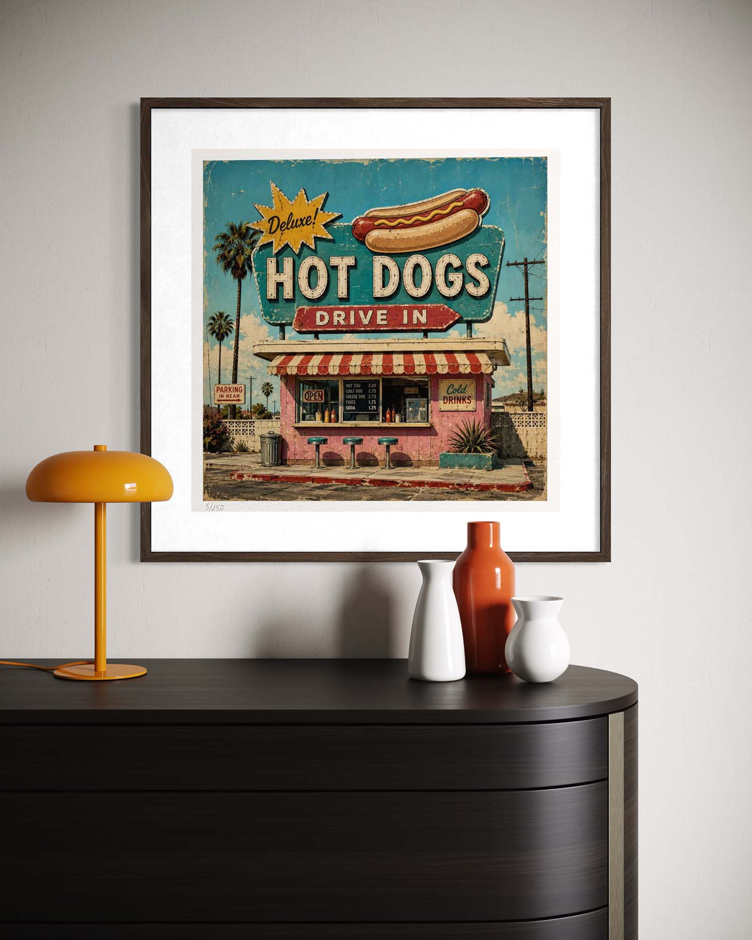 A framed retro-style hot dog restaurant poster hanging on a light gray wall above a dark wooden console table. The poster features a pink diner, a hot dog illustration, and bold text reading 'Deluxe Hot Dogs Drive In.