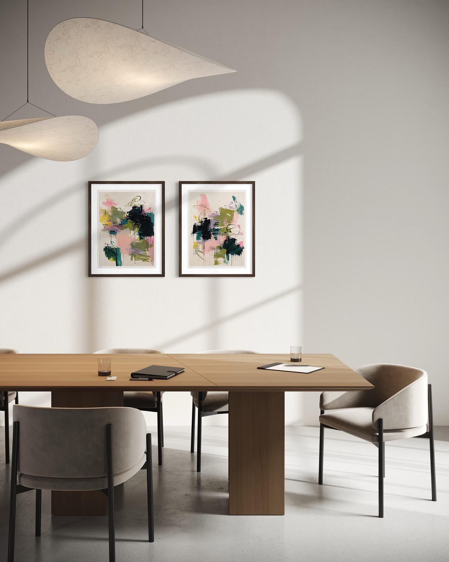 Two framed abstract art prints with black frames hang on a light gray wall above a wooden dining table and gray upholstered chairs in a modern room with pendant lights casting shadows.