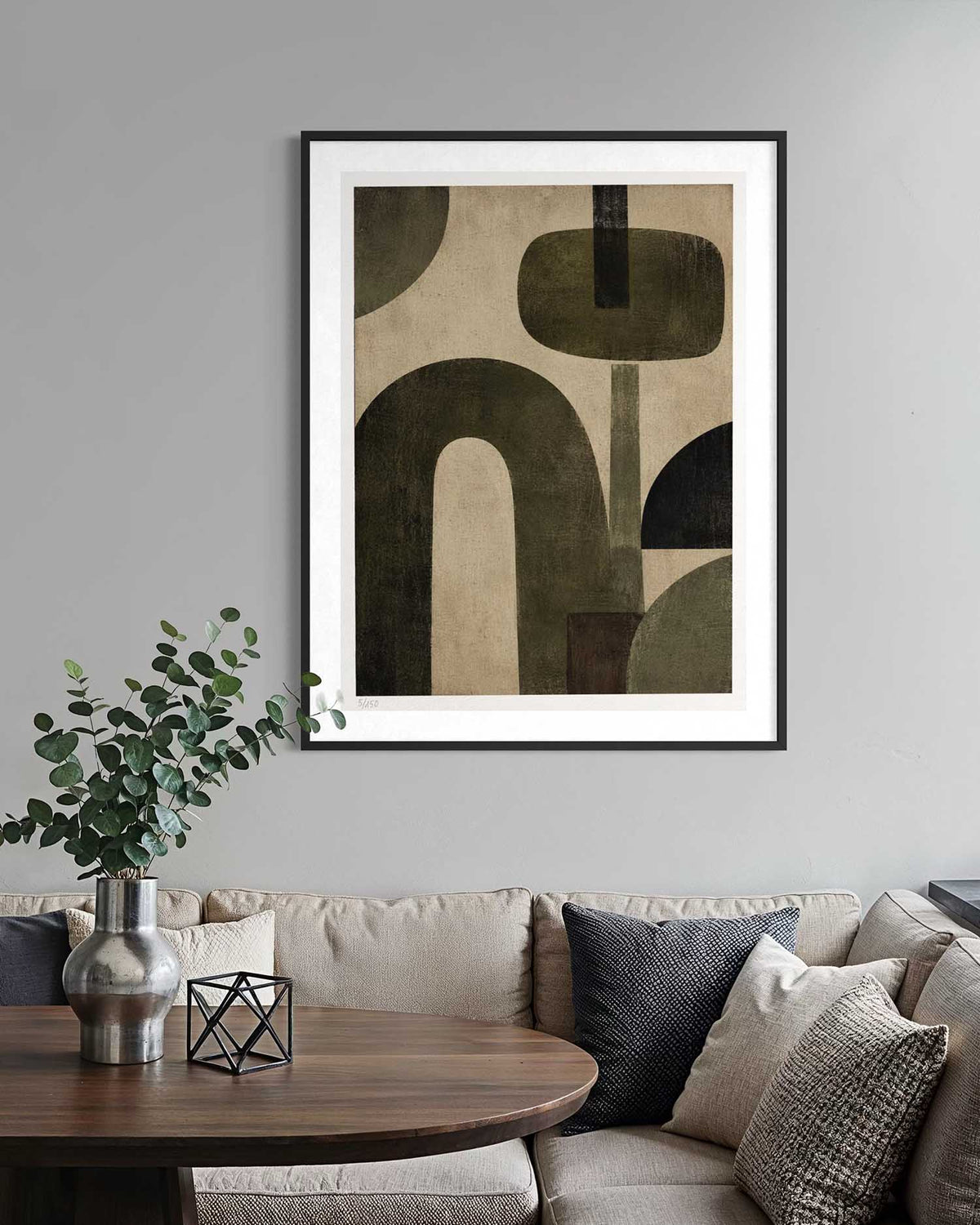 A framed abstract art print with geometric shapes in olive green, dark brown, and beige tones, displayed on a light gray wall above a beige sofa with decorative pillows and a wooden table in the foreground.