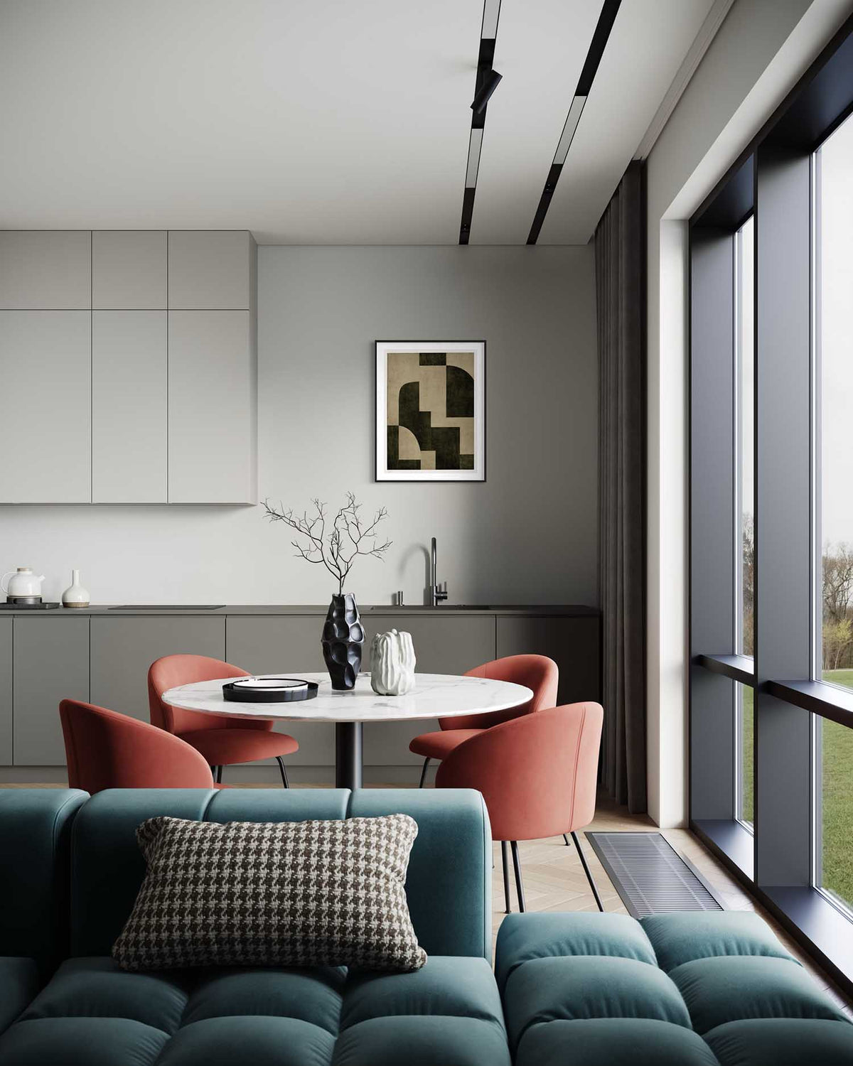 A modern interior scene showing a teal tufted sofa in the foreground, a round dining table with four coral-red chairs, a gray kitchen cabinetry wall, and a large window with dark gray framing. A framed abstract print hangs on the wall.