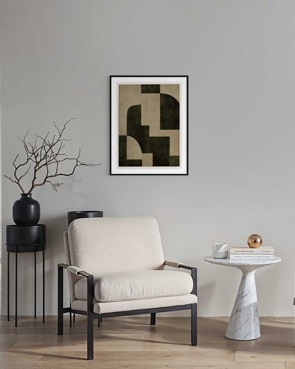 A framed abstract geometric art print with black and beige blocks hangs on a light gray wall above a cream upholstered armchair in a modern living room setting.