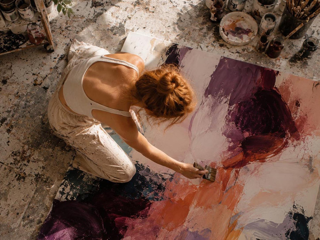 An overhead view of a woman painting on a large canvas in a studio. She is kneeling and wearing a white tank top and light-colored pants, using a brush to apply paint.