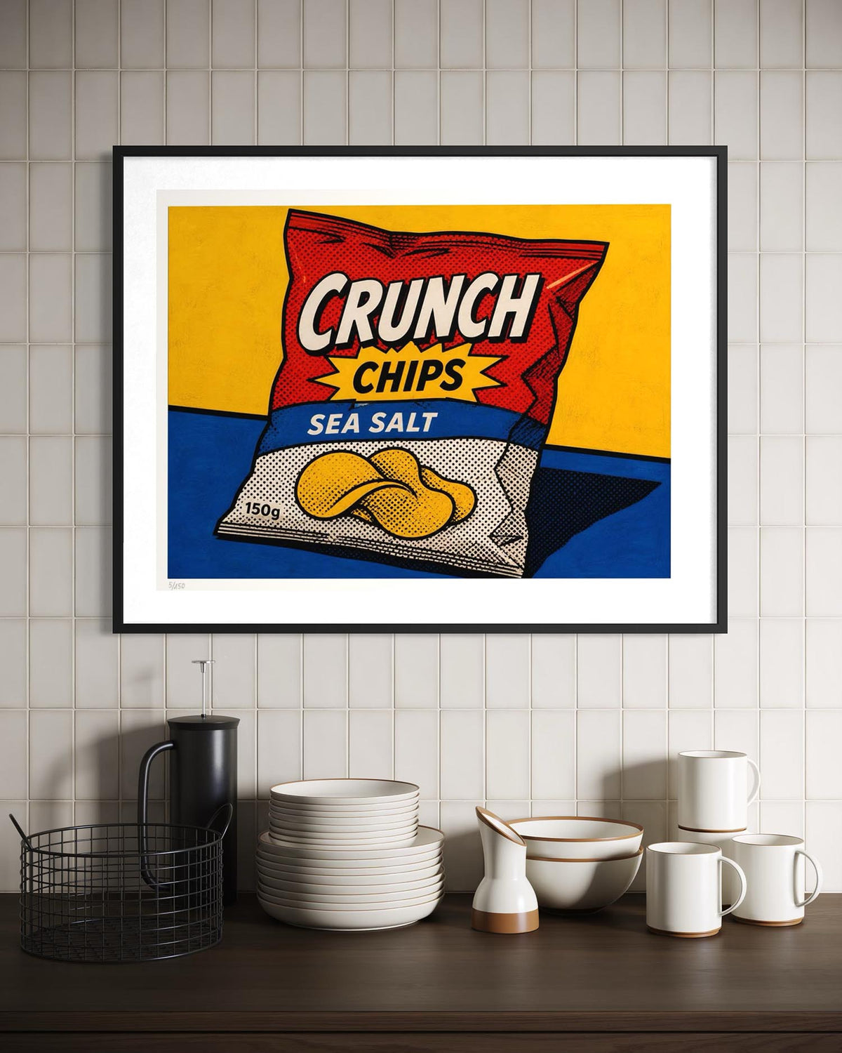 A framed pop-art style print of a Crunch Chips Sea Salt bag hanging on a white tiled wall above a dark wooden shelf with white dishes and mugs.