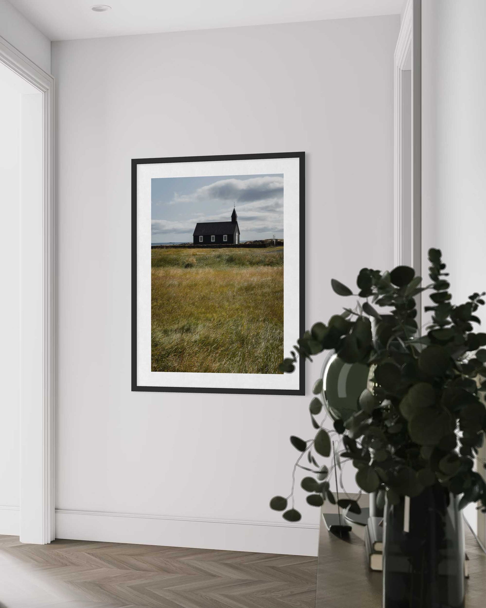 Framed photograph of a house in a field on a white wall with a plant in the foreground.