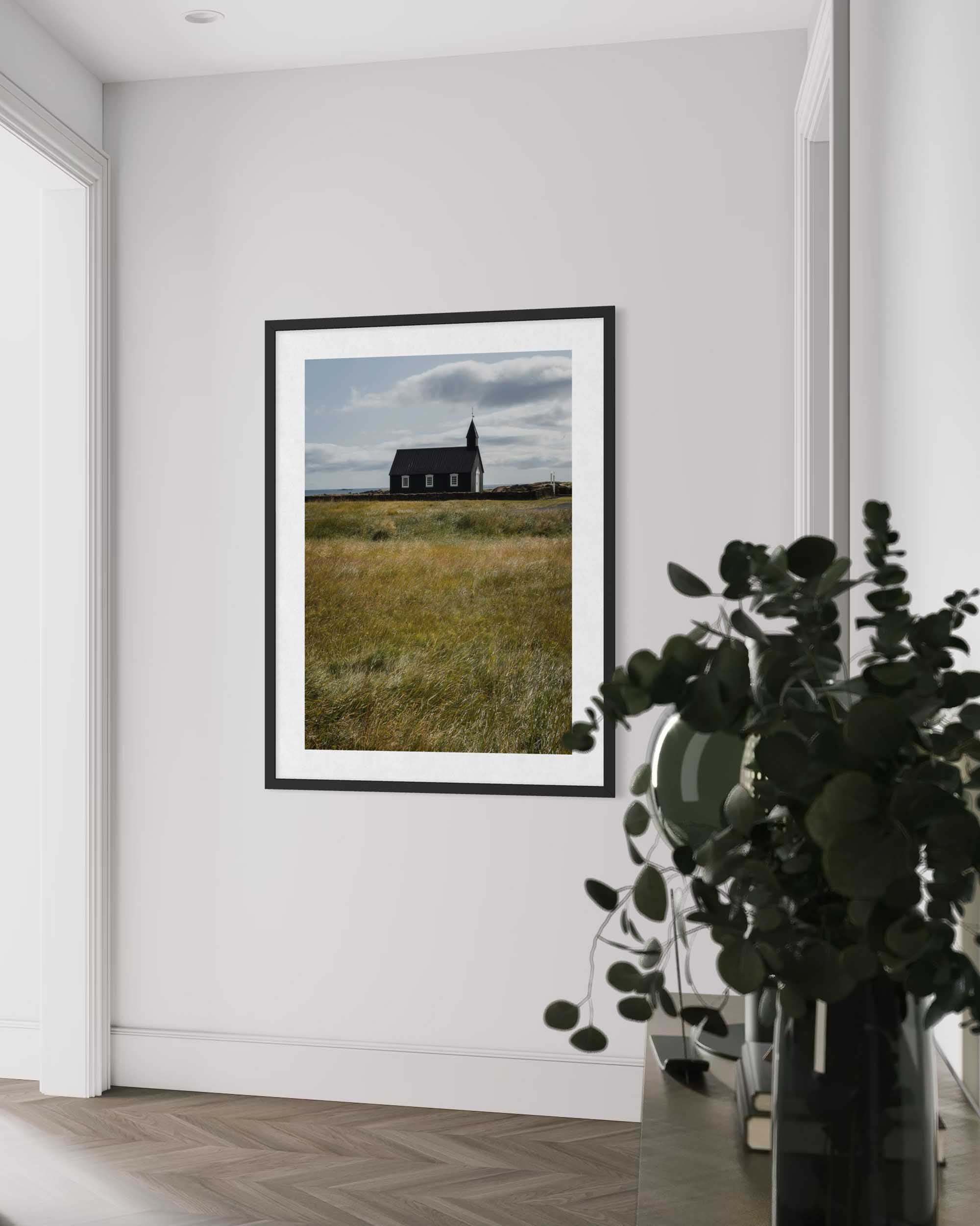 Framed photograph of a house in a field on a white wall with a plant in the foreground.