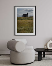 Framed photograph of a church in a field above a modern chair in a room.