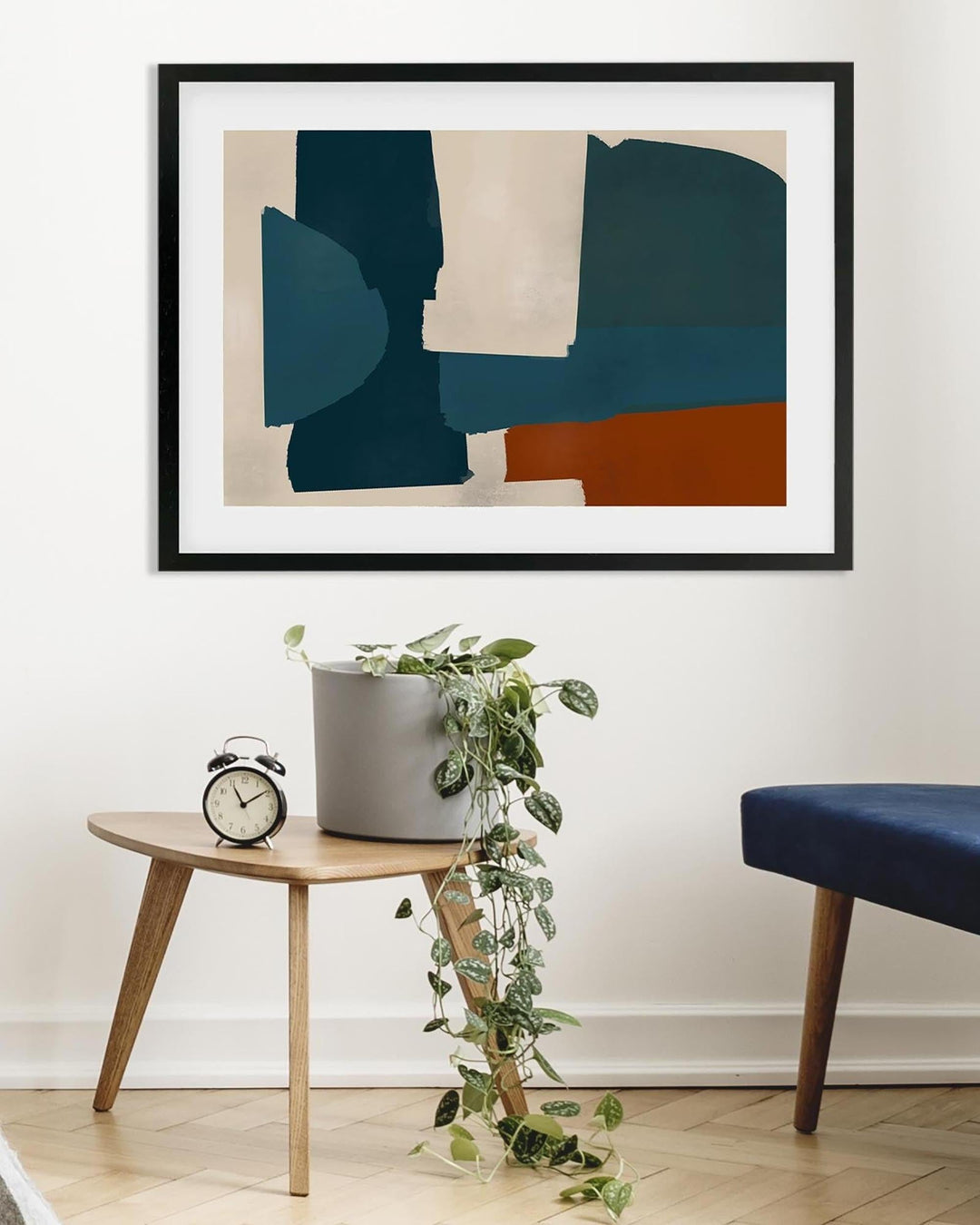 Abstract art piece in a black frame on a wall above a wooden side table with a plant and clock.