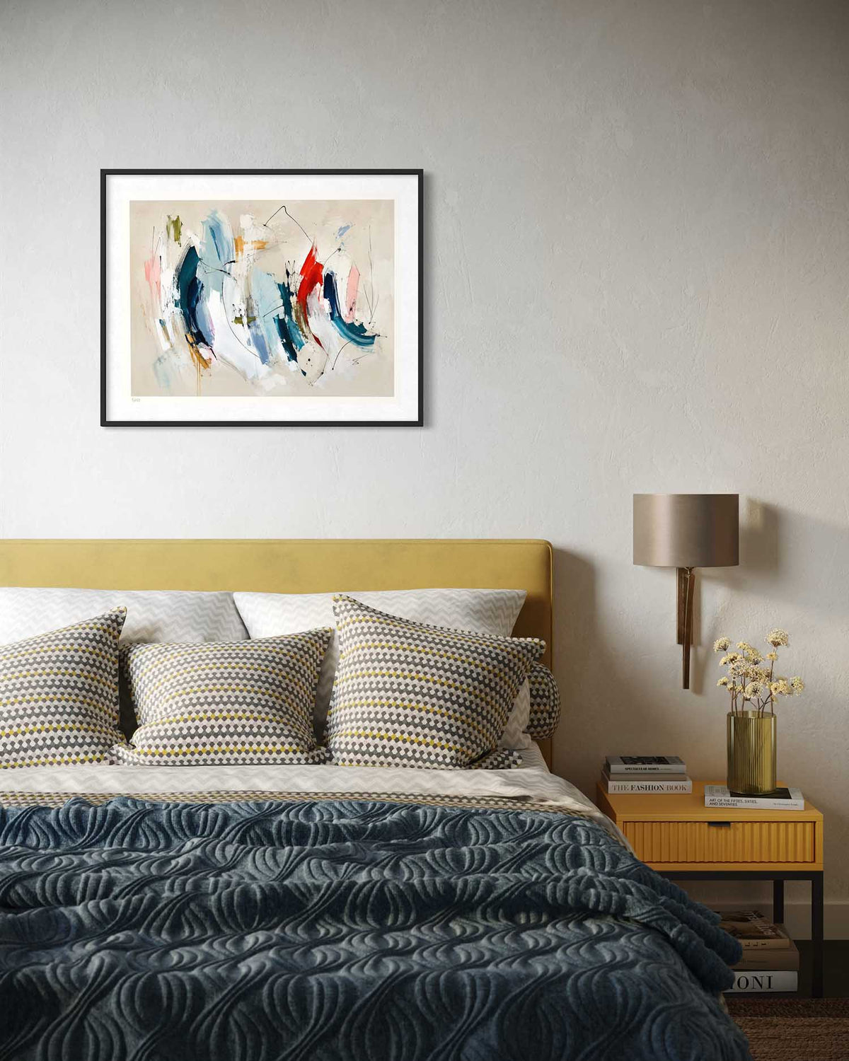 An abstract framed wall art print hangs above a bed in a styled bedroom. The artwork features expressive brushstrokes in blue, white, red, and beige on a light background, enclosed in a thin black frame.