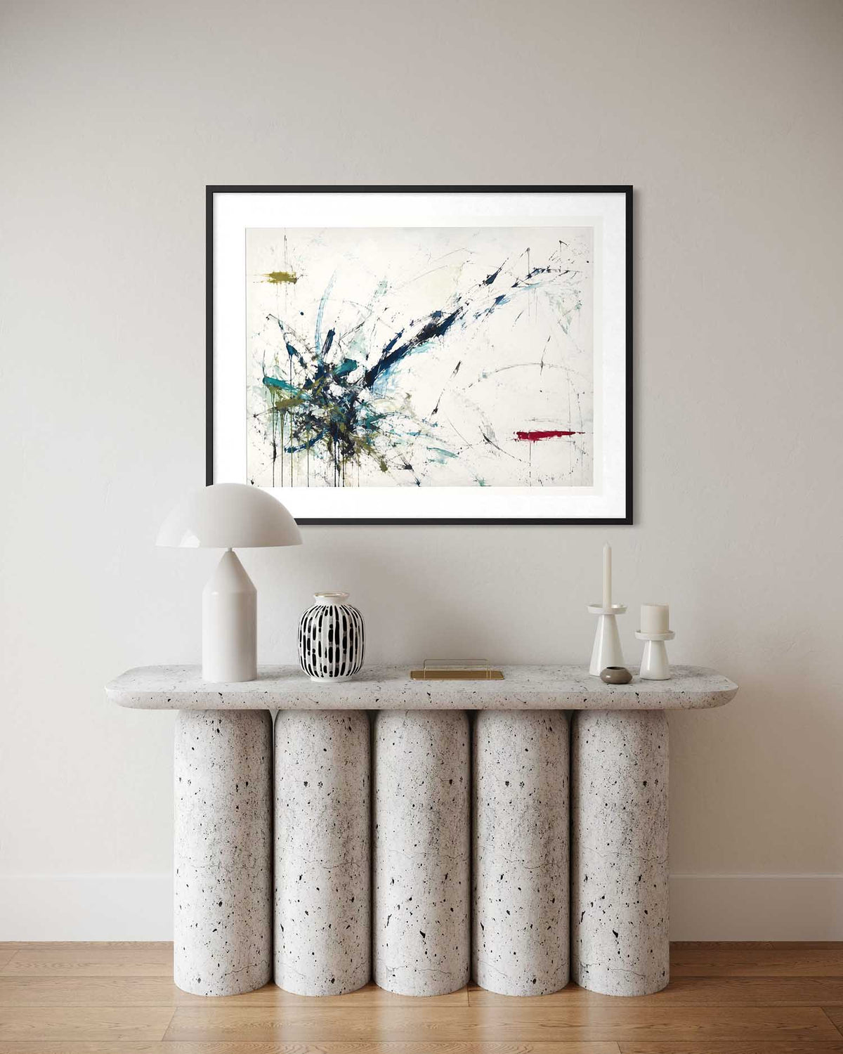 A framed abstract art print with blue, black, white, and red paint strokes hangs on a beige wall above a modern console table with cylindrical stone legs and white decor.