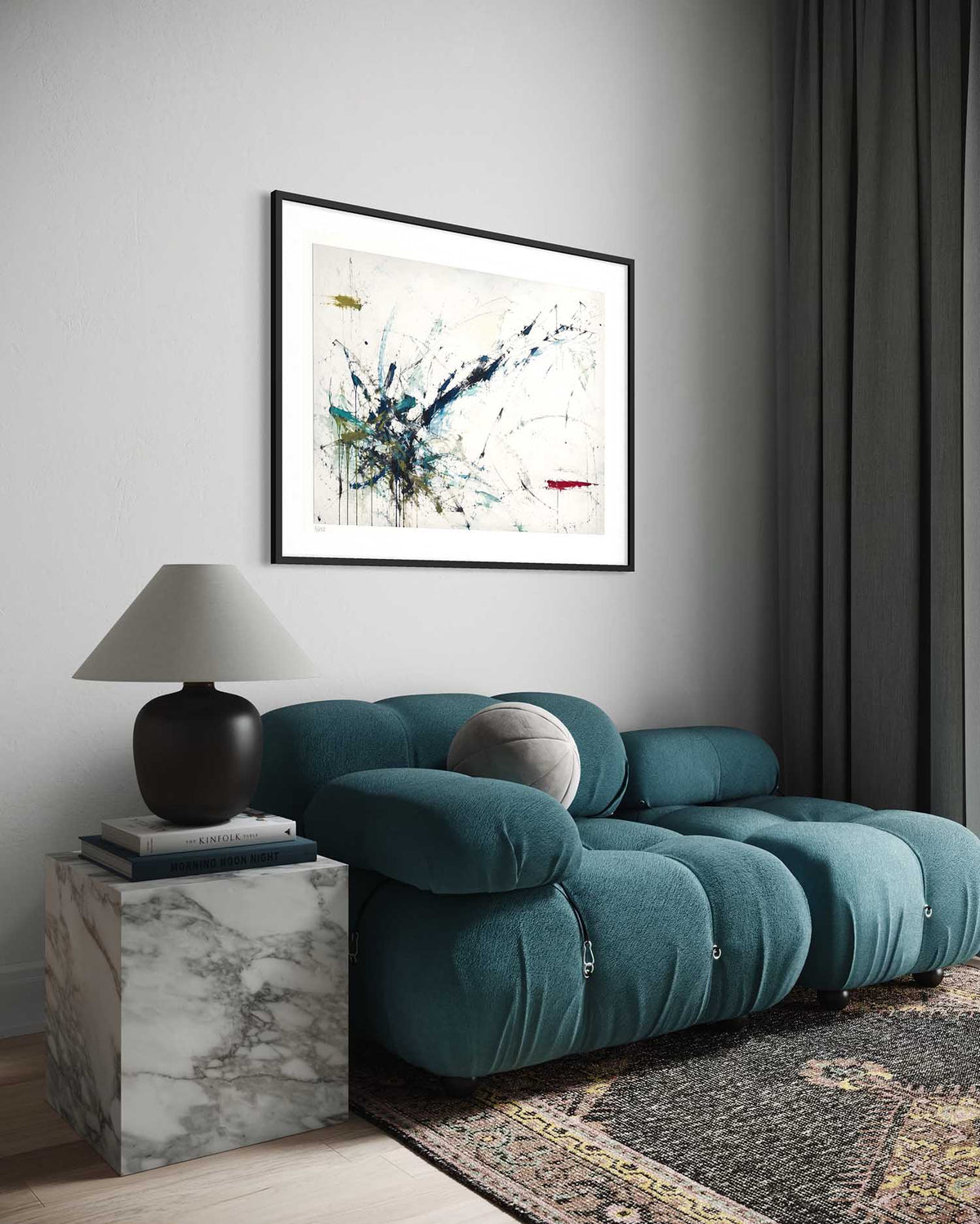 A framed abstract art print hangs on a light gray wall above a teal tufted sofa in a modern living room setting. The print features blue, black, and white paint splatters on a cream background with a thin black frame.