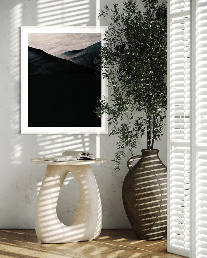 Decorative setup with a framed artwork, white side table, and vase against a wall with sunlight filtering through blinds.