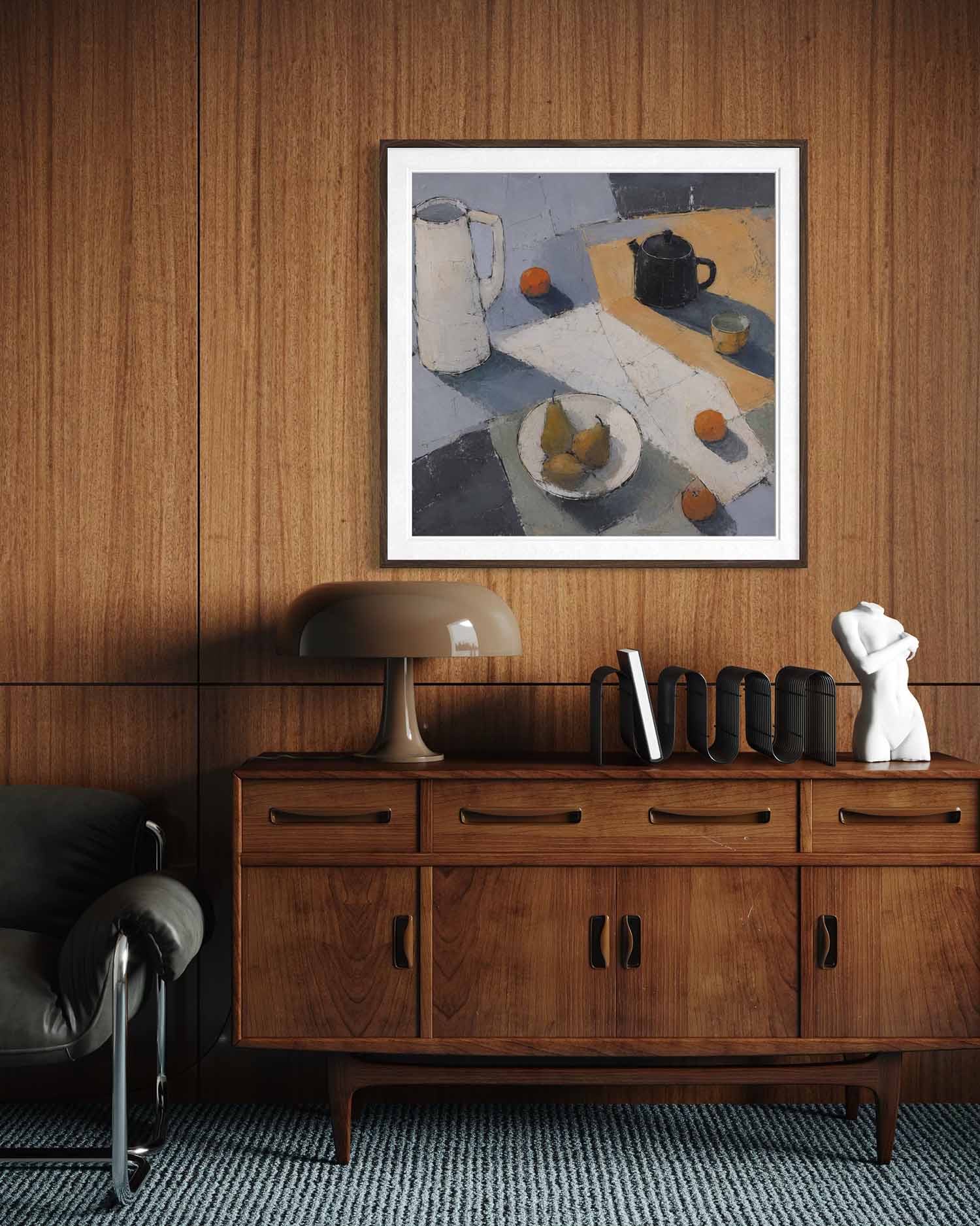 A framed still life painting with muted blue, gray, beige, and orange tones hangs on a wood-paneled wall above a mid-century modern wooden credenza with a lamp and decorative objects.