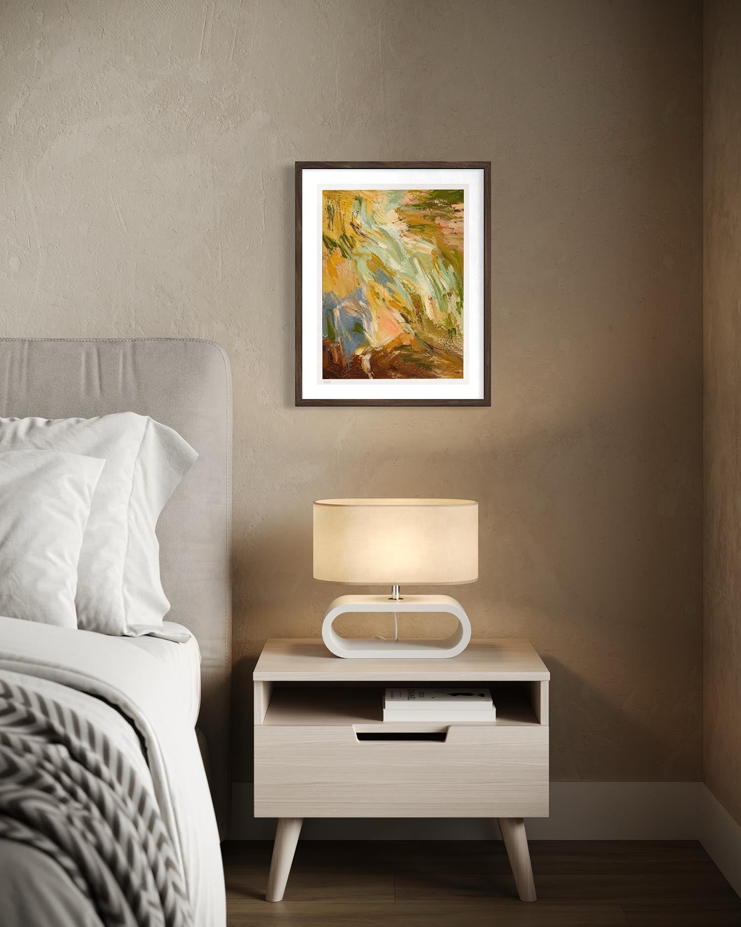 A framed abstract landscape art print with warm earthy tones of green, yellow, and orange, displayed on a textured taupe wall above a light wood nightstand with a lit lamp and white bedding.